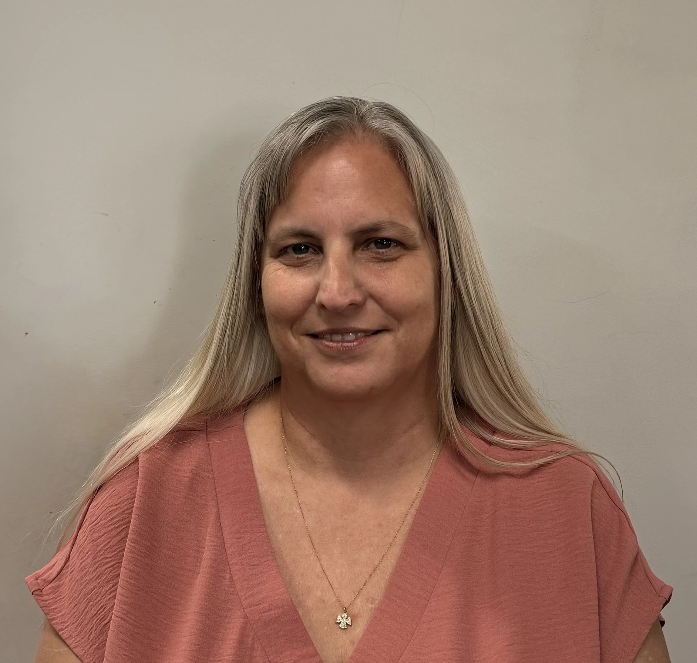 A middle-aged woman with long blonde hair wearing a salmon-colored top and a small gold necklace with a charm, smiling softly at the camera against a plain light-colored background.