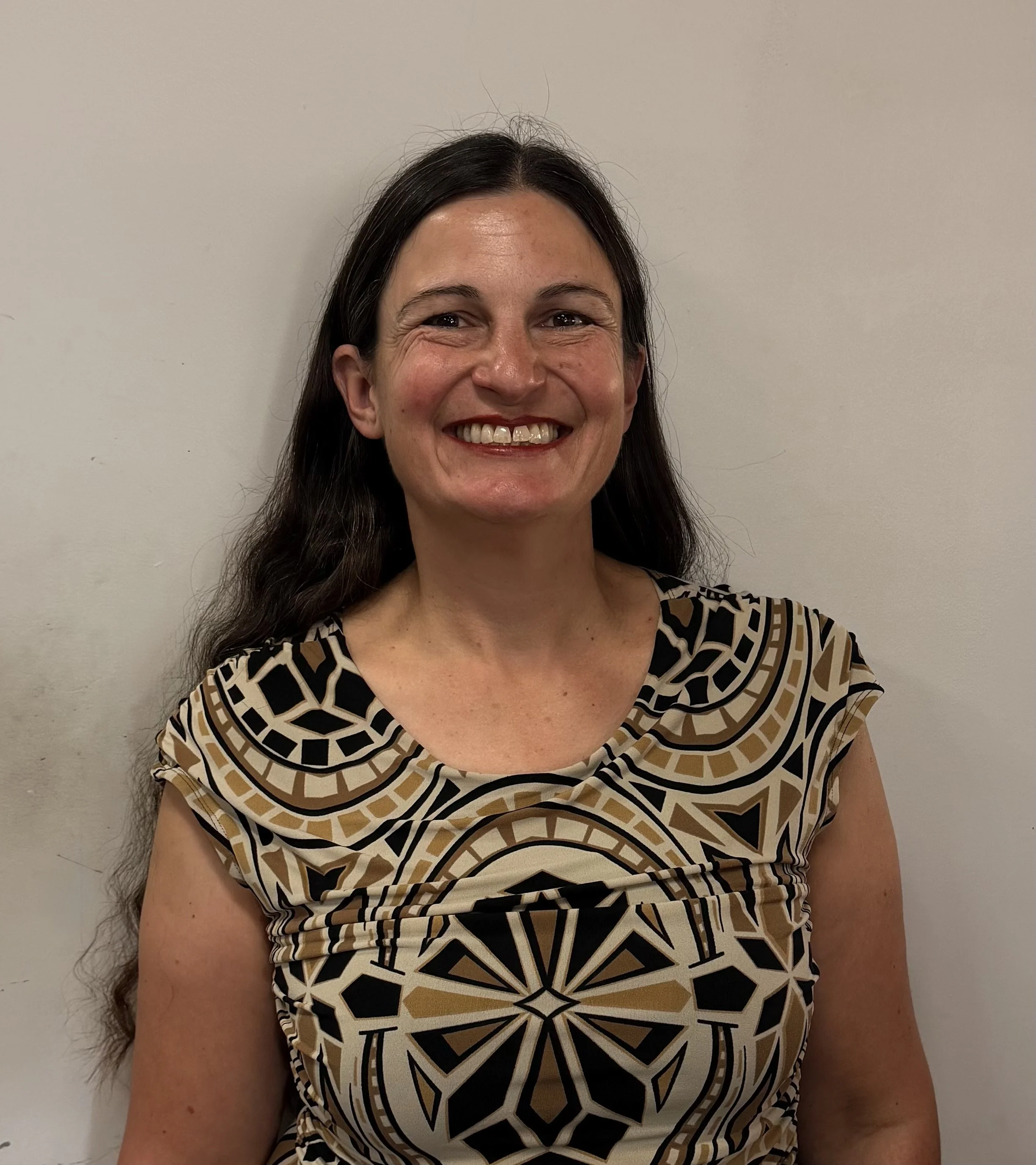 A woman with long dark hair smiling, wearing a patterned black, beige, and white top, standing against a plain light-colored wall.