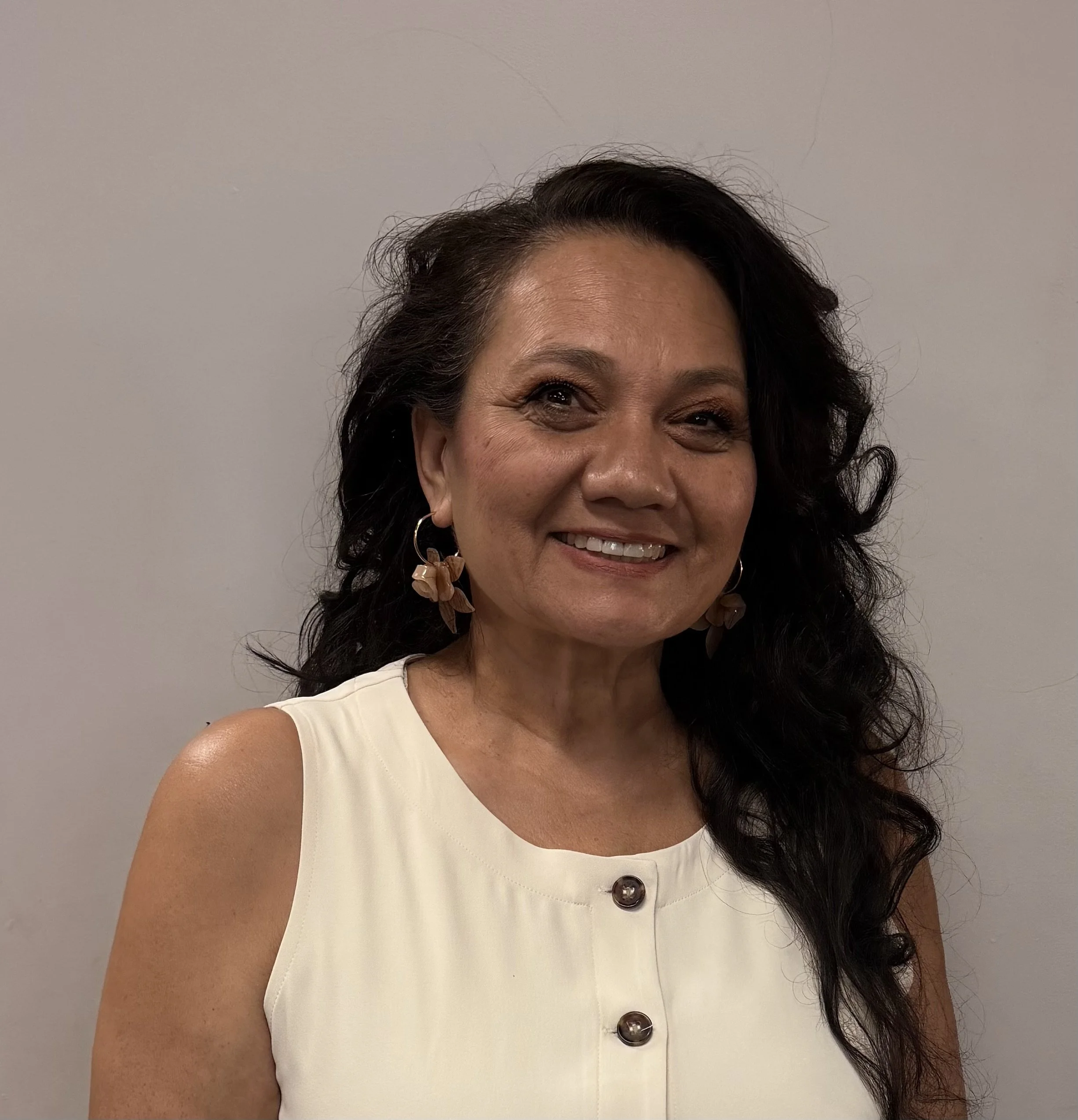 A smiling woman with dark, curly hair wearing a sleeveless white top and floral earrings, standing against a plain light background.