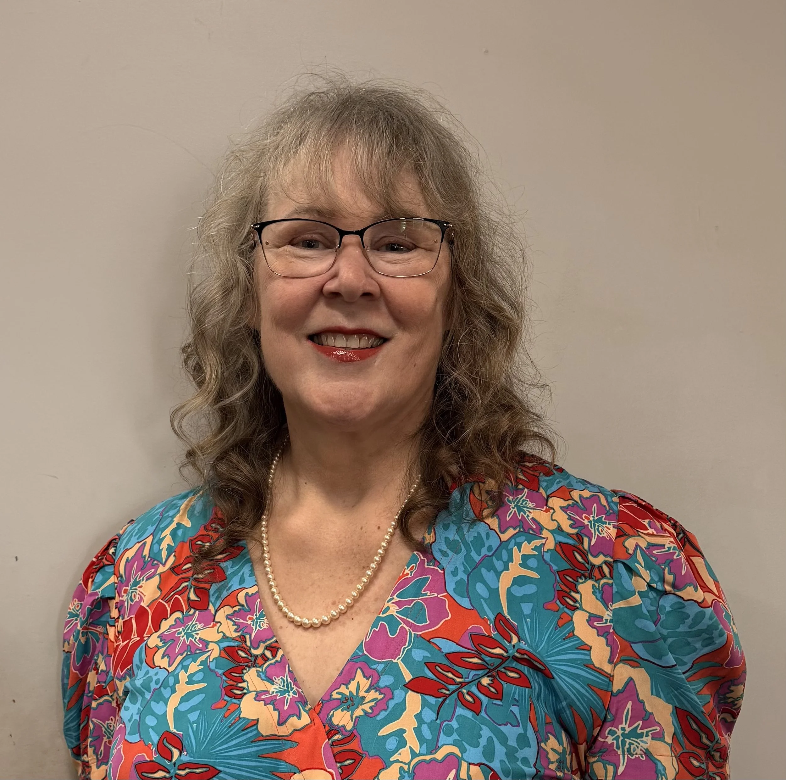A middle-aged woman with curly gray hair, wearing glasses, a vibrant floral dress, and a pearl necklace, smiling against a plain background.