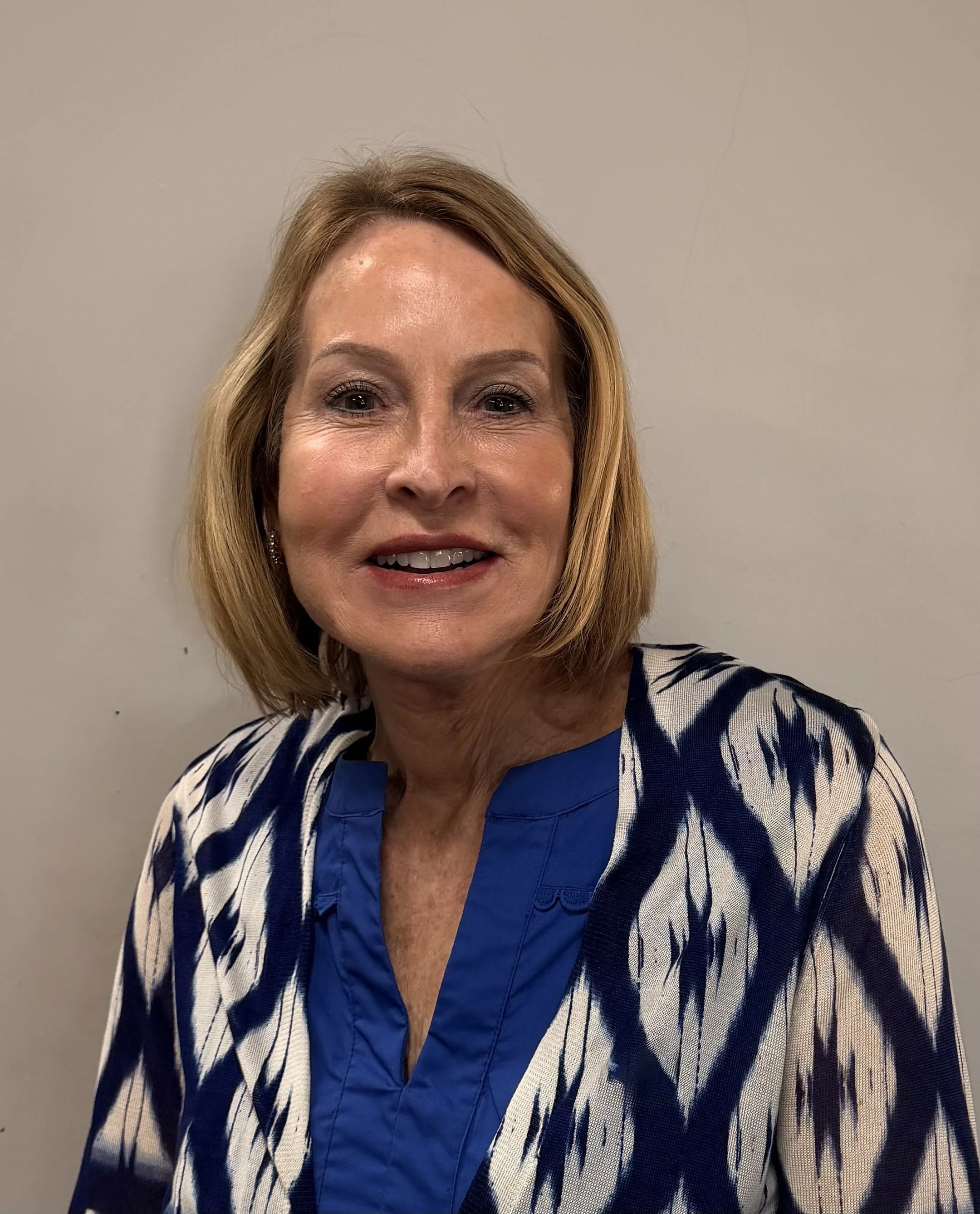 A woman with shoulder-length blonde hair smiling, wearing a blue and white patterned blouse, standing against a plain gray wall.