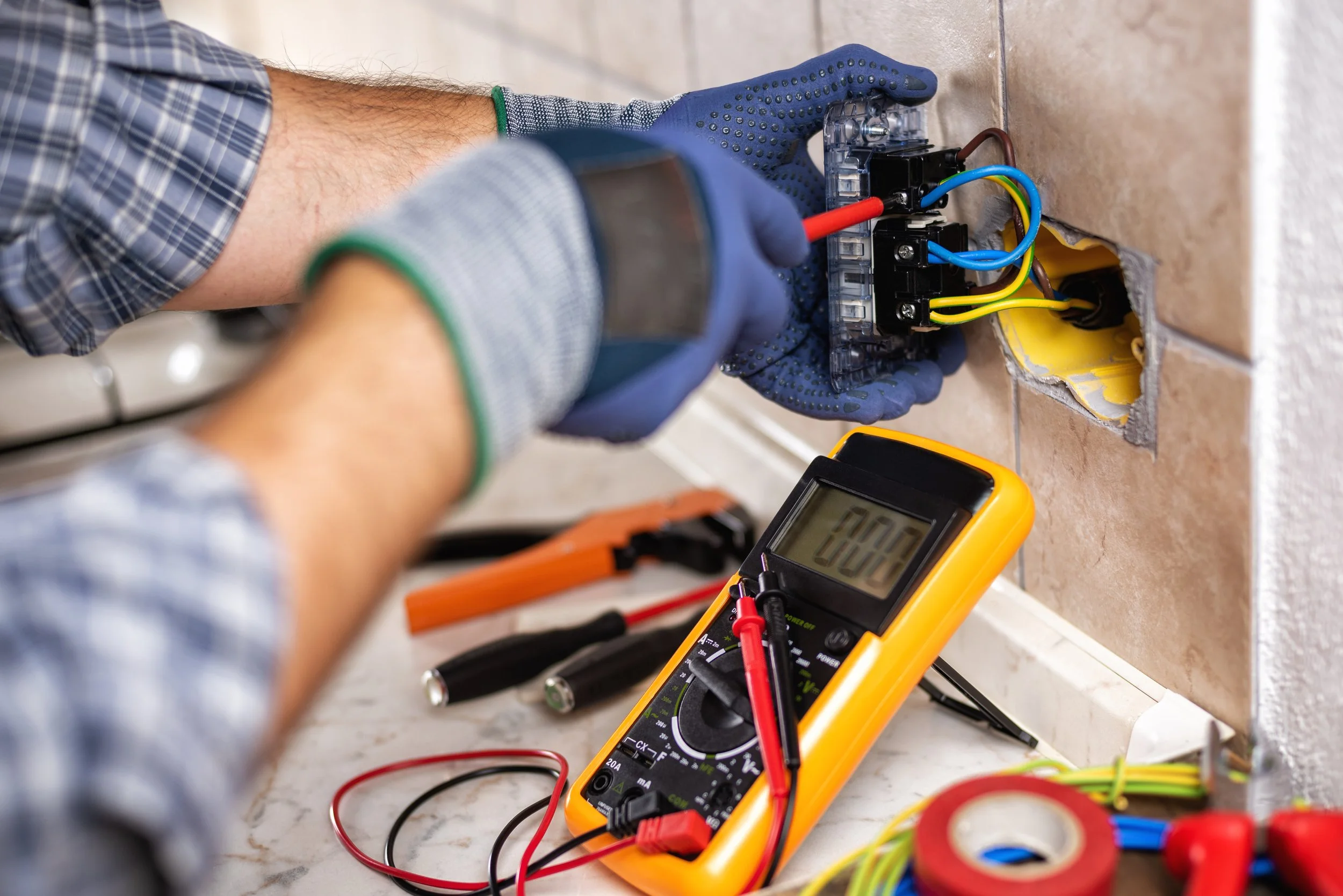 A person wearing a plaid shirt and blue gloves is using a screwdriver to work on an electrical outlet with various colored wires. A digital multimeter is set to 200, with red and black probes, and is placed on the countertop near the person. Tools and wires are scattered around on the surface.