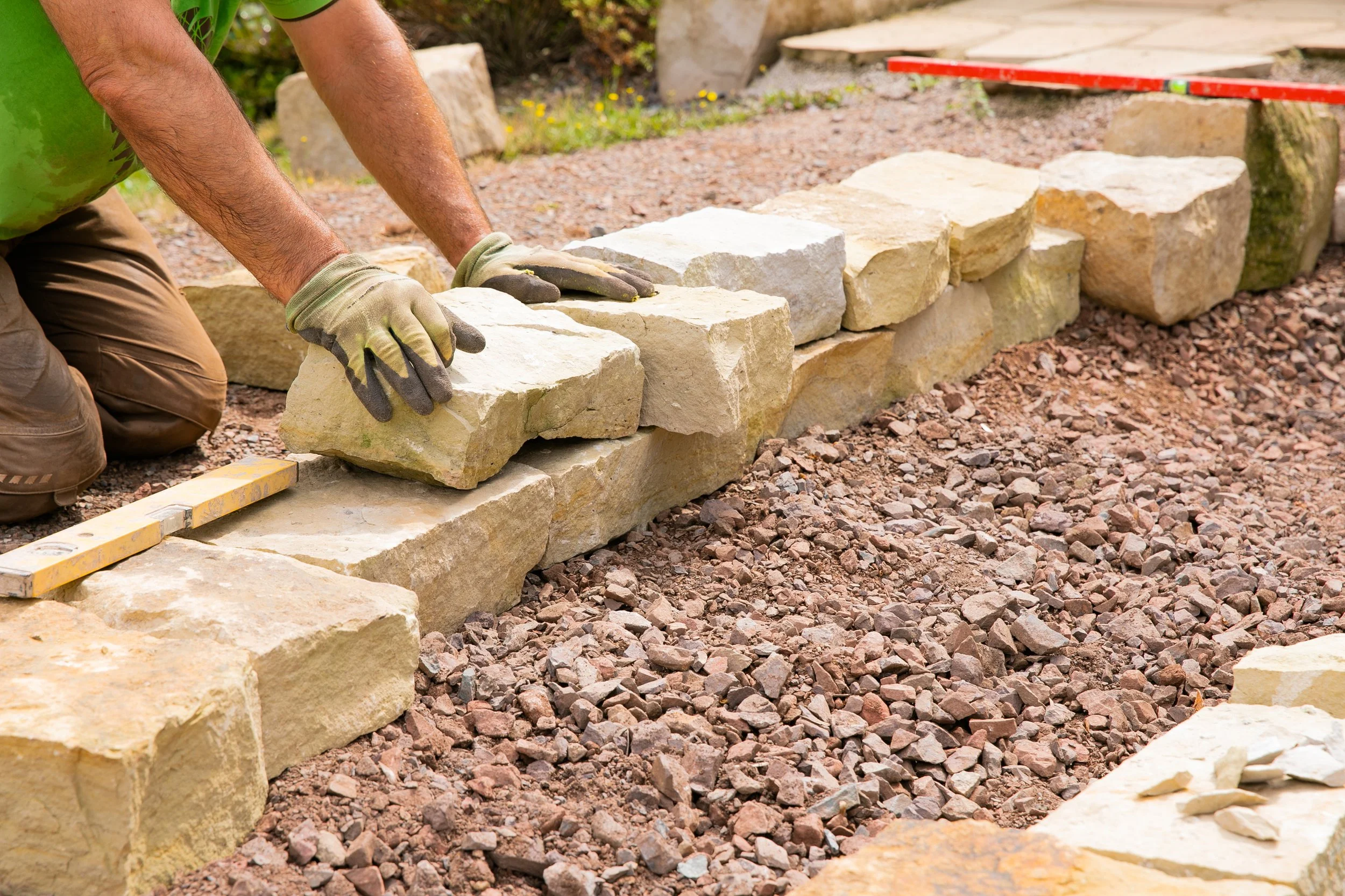 A person laying large beige and light yellow stones to build a stone border on a gravel surface, wearing gloves and kneeling.