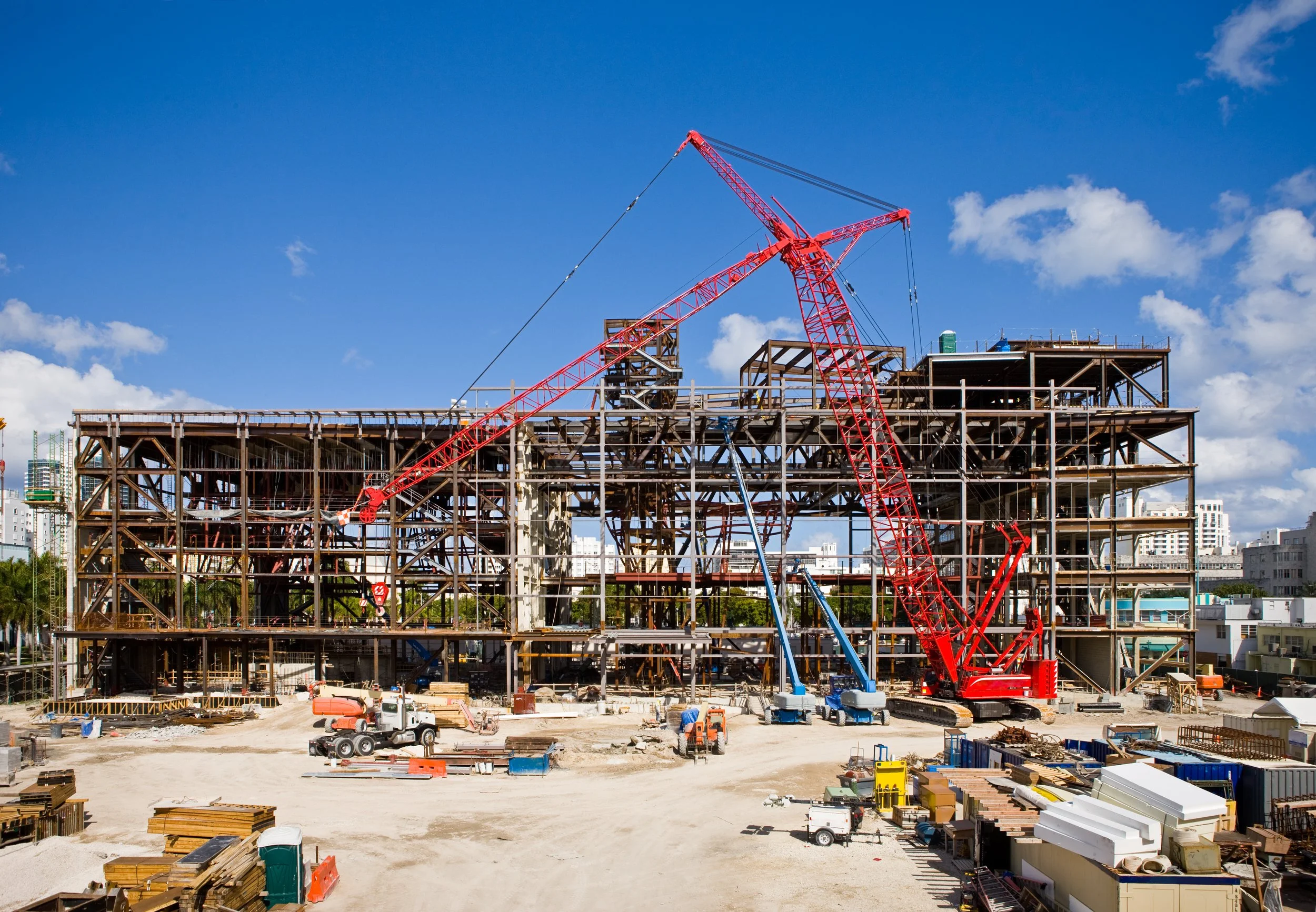 Construction site with a large red crane, unfinished steel framework, construction vehicles, and building materials against a blue sky with scattered clouds.