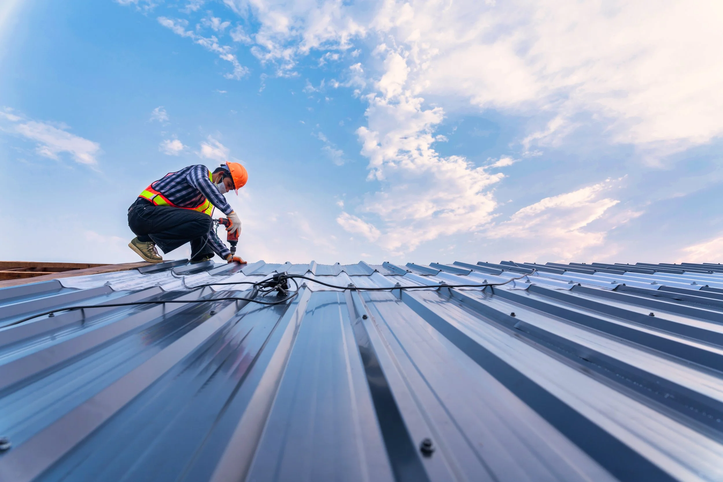 A worker wearing safety gear and a high-visibility vest installing or repairing a metal roof.