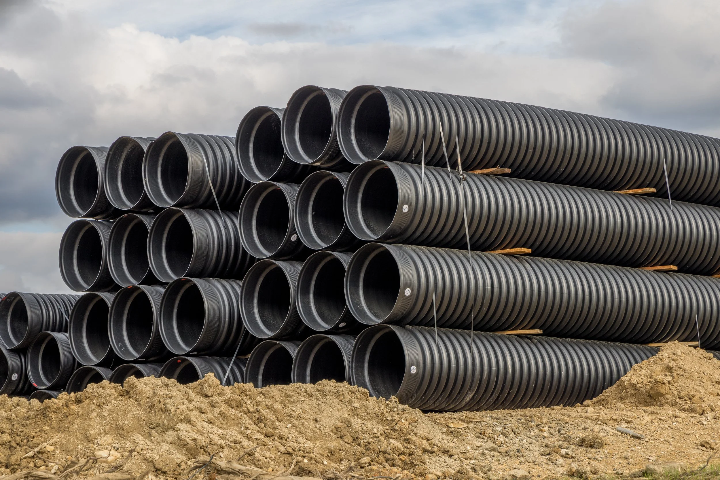 Stacks of large black corrugated plastic pipes on a construction site with dirt ground and a cloudy sky.