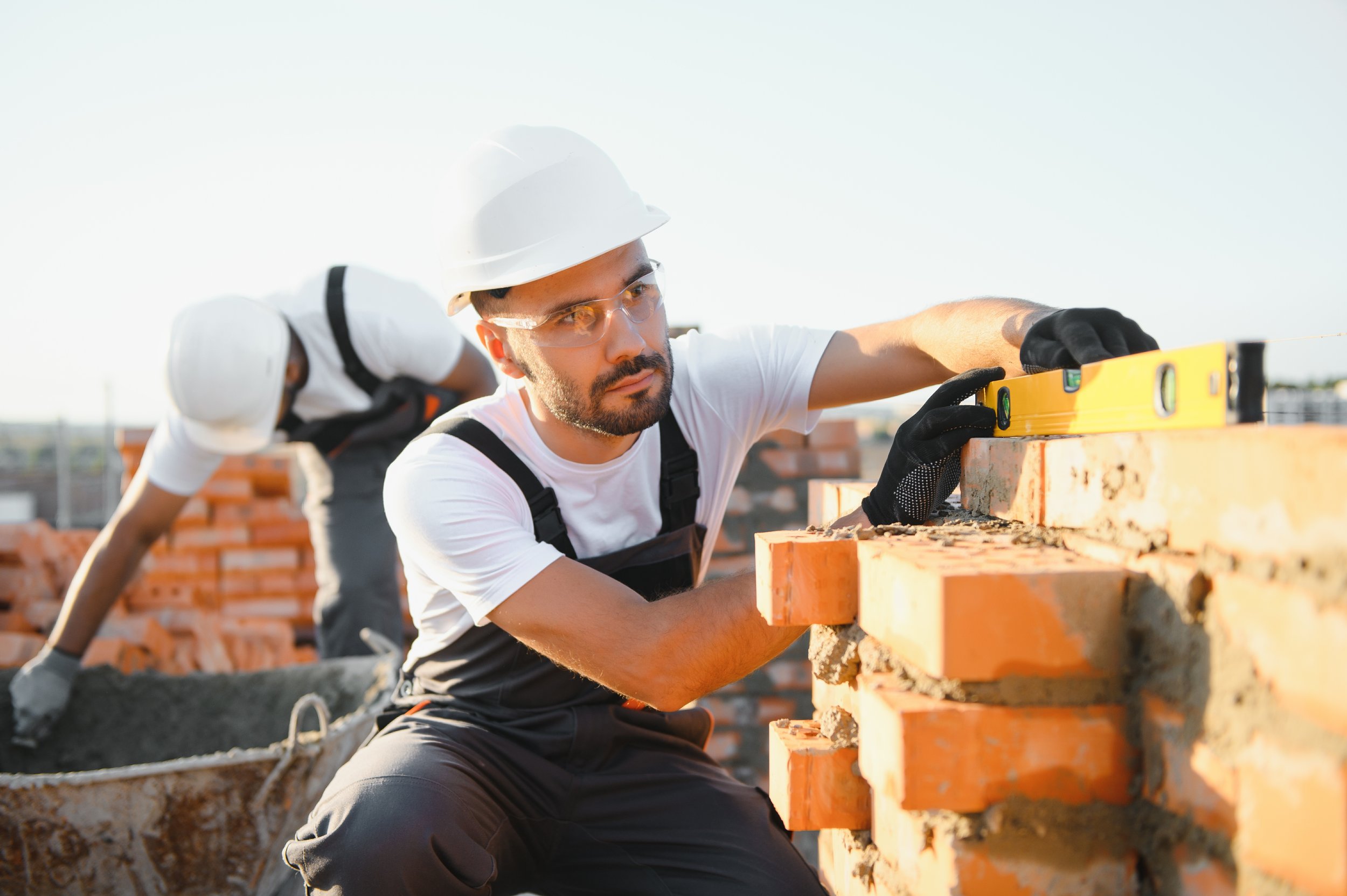 Two construction workers, wearing white helmets and safety goggles, are building a brick wall outdoors during daytime. One worker is measuring with a level tool on the bricks.