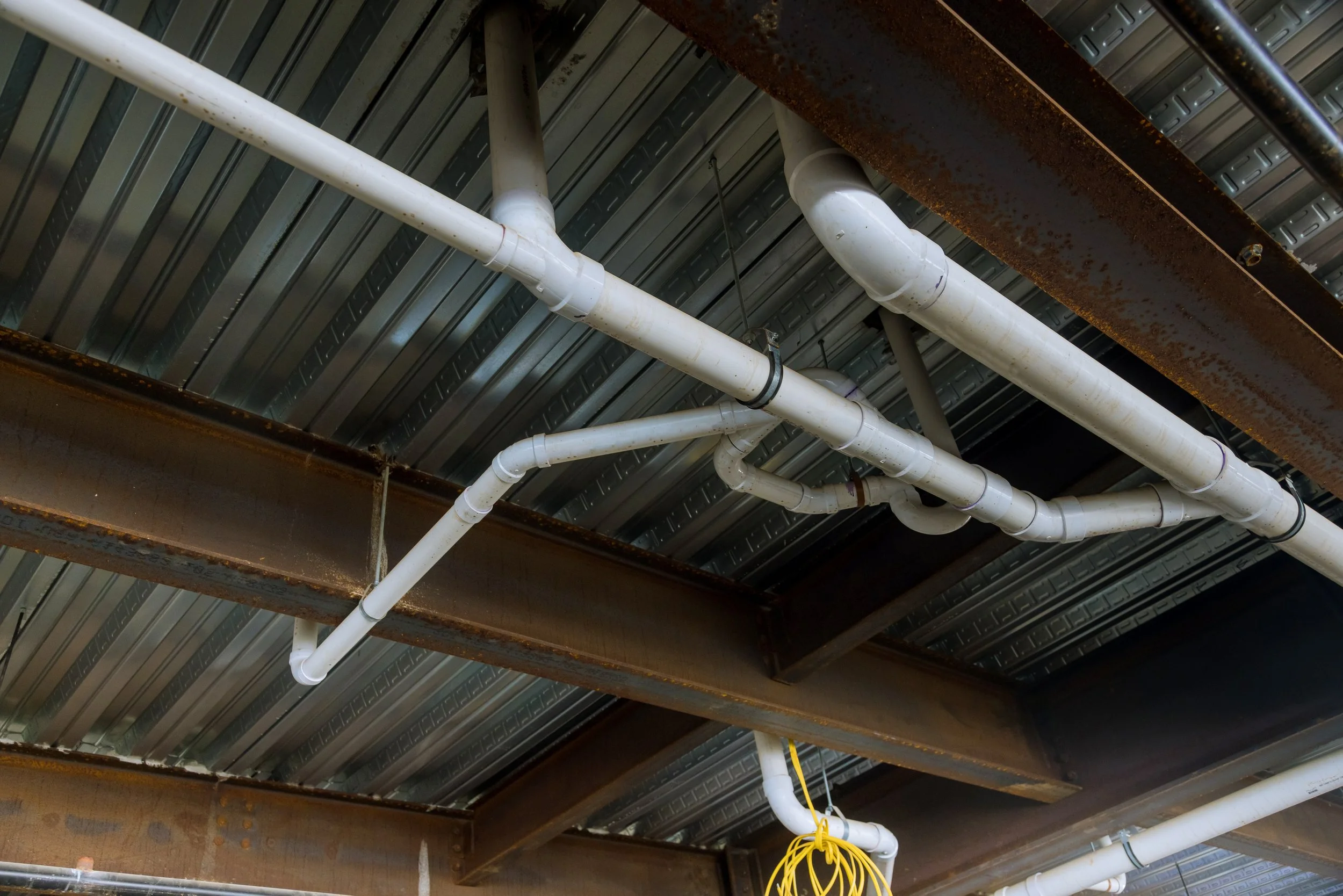 Ceiling with white PVC piping, rusty steel beams, and corrugated metal roof.
