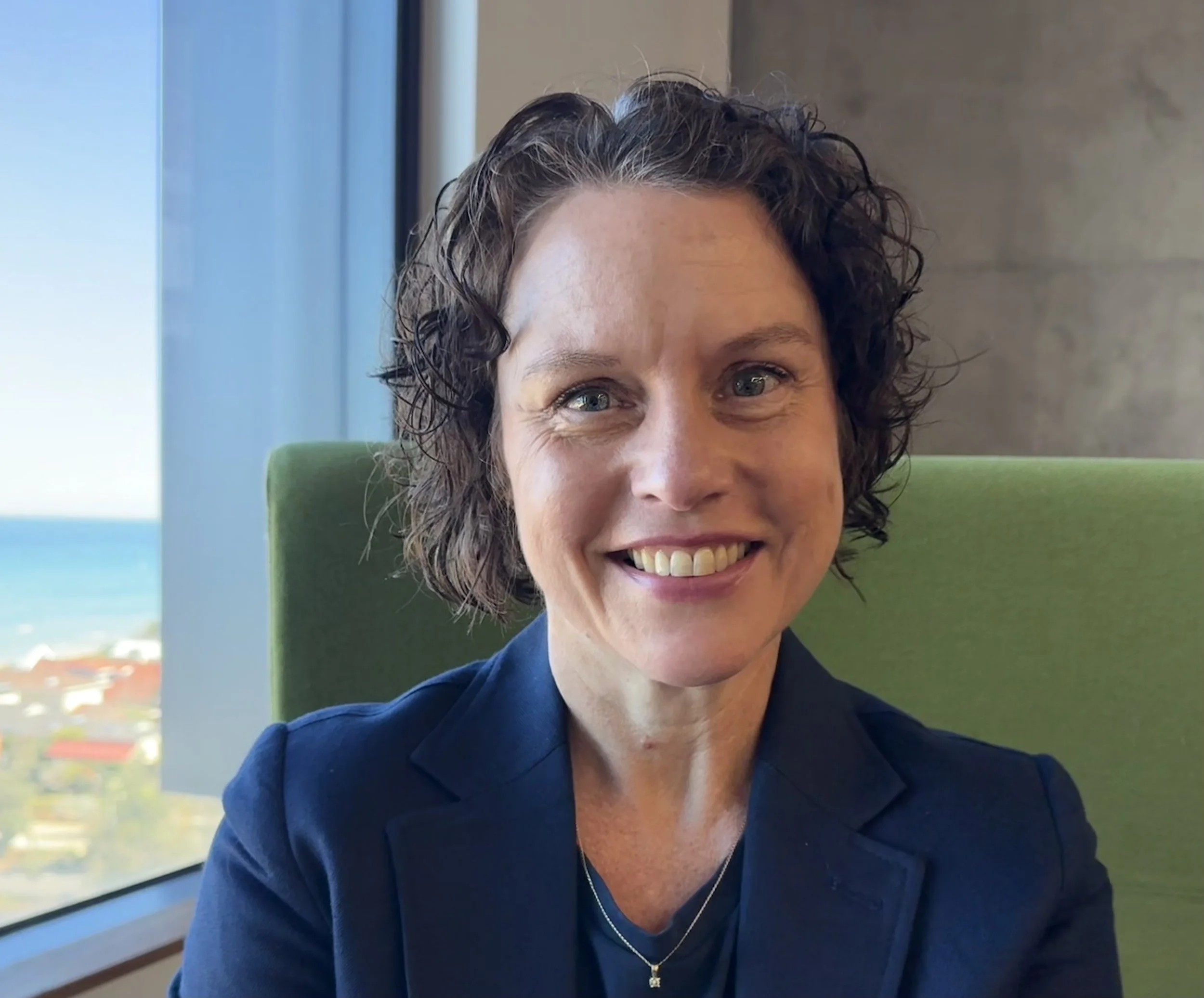 A woman with shoulder-length curly brown hair smiling, wearing a navy blazer and a delicate necklace, sitting in front of a window with a view of the city and ocean in the background.
