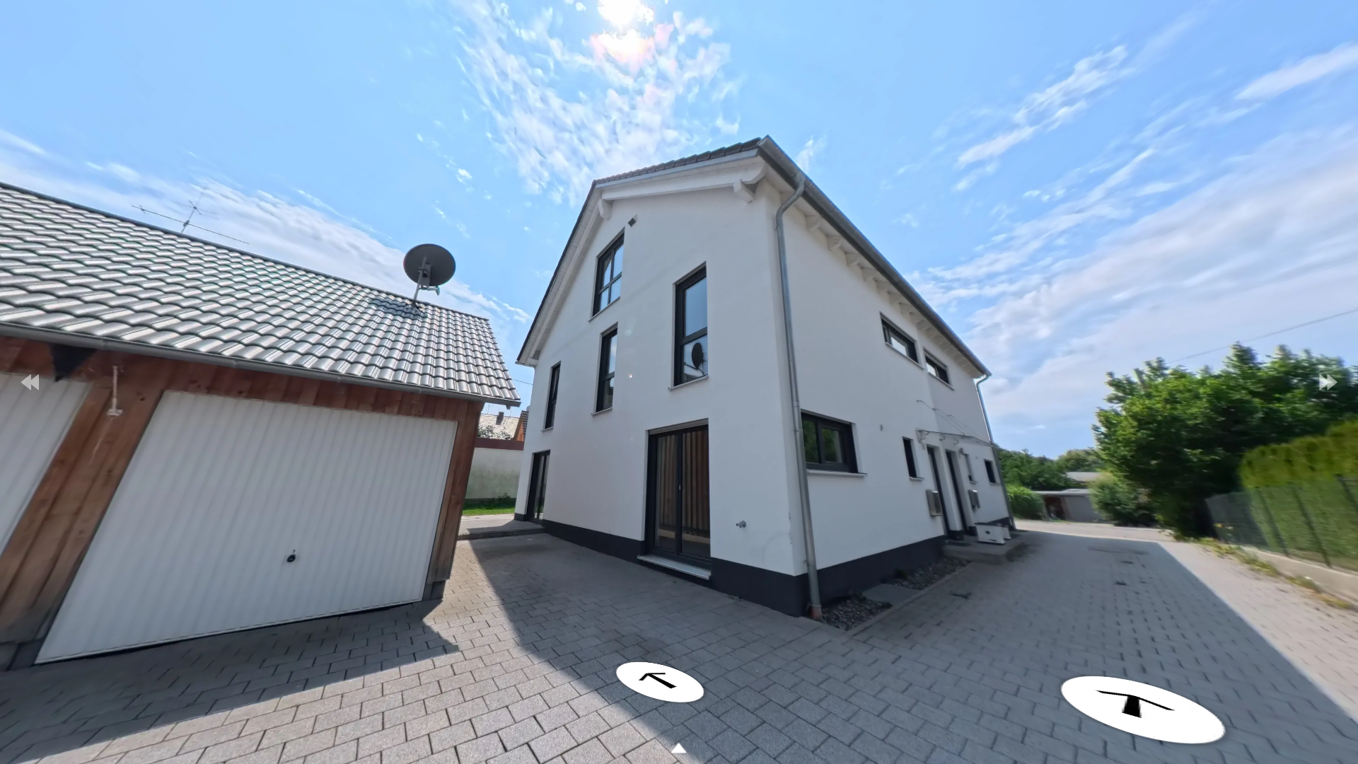 Ein modernes, weißes Mehrfamilienhaus mit mehreren Fenstern und einem Satteldach, neben einem kleineren Loch für eine Garage mit grauem LOGO-Gartenhäuschen auf einem gepflasterten Hof und einer grünen Baumlinie im Hintergrund bei sonnigem Himmel.