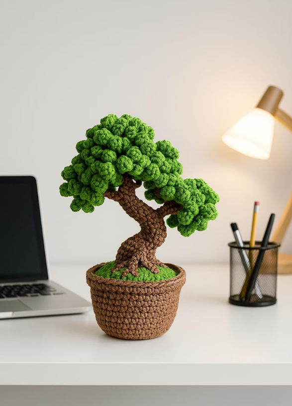 A crocheted potted bonsai tree with green leaves and a brown trunk on a white desk, with a laptop, a pen holder, and a desk lamp in the background.