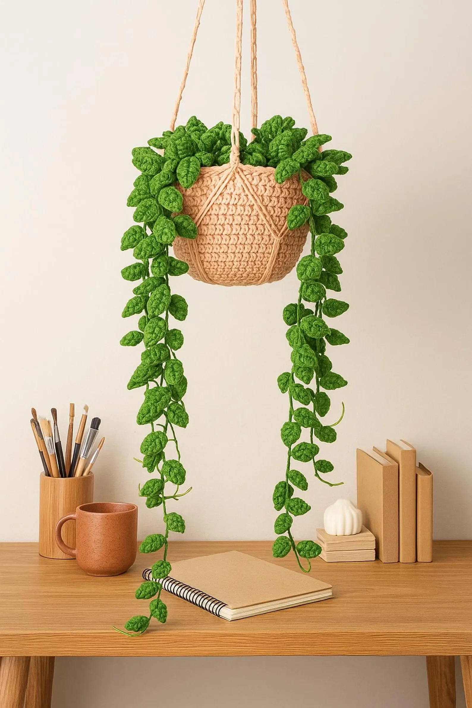 A hanging pink macrame plant holder with green faux trailing leaves, suspended above a wooden desk. The desk has a pencil holder with pens, a ceramic mug, a spiral notebook resting on a closed tan notebook, some stacked books, and a decorative pumpkin-shaped object.