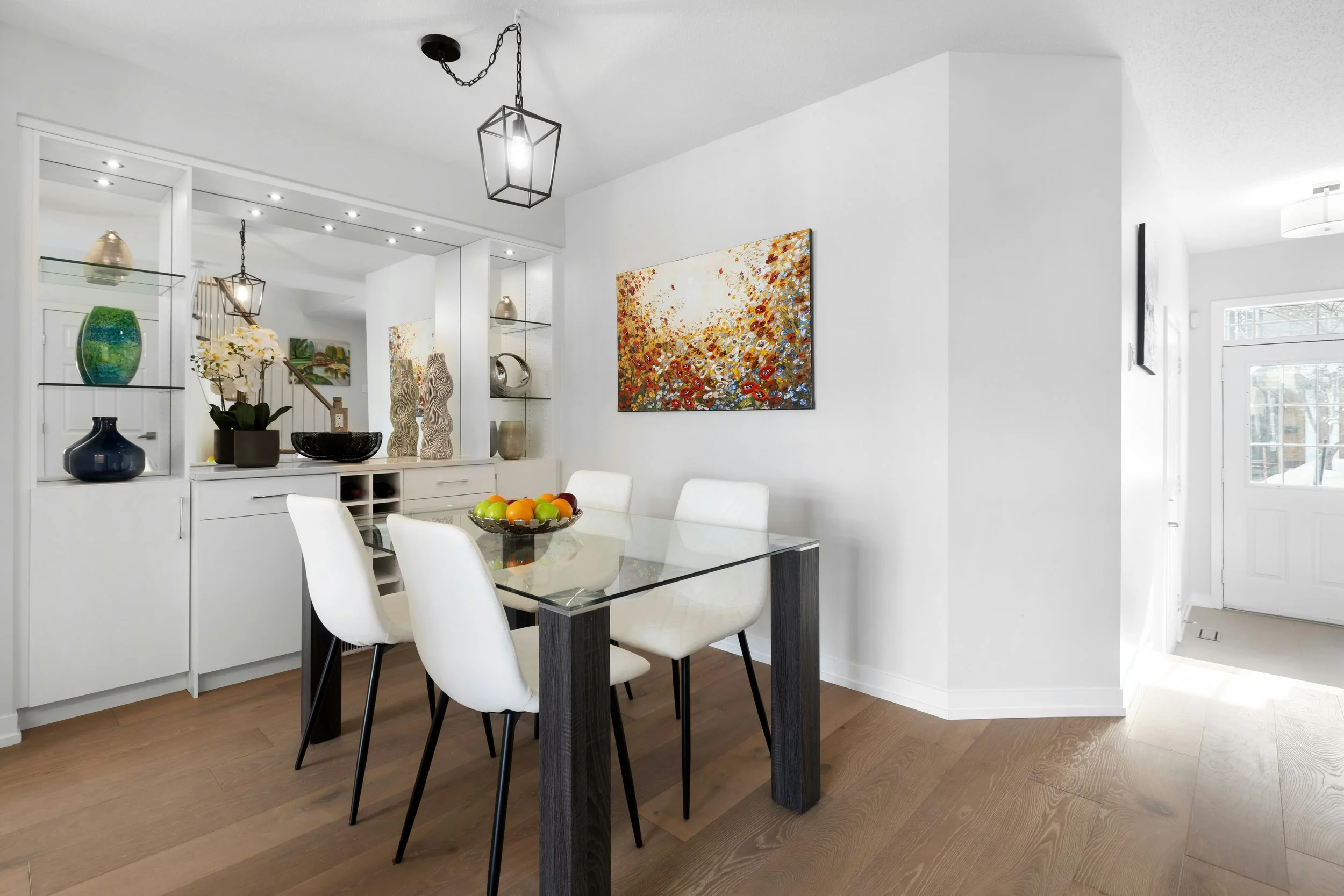 A dining room with a glass-top table, white chairs, and decorative vases and artwork, with a white wall and wooden floor.