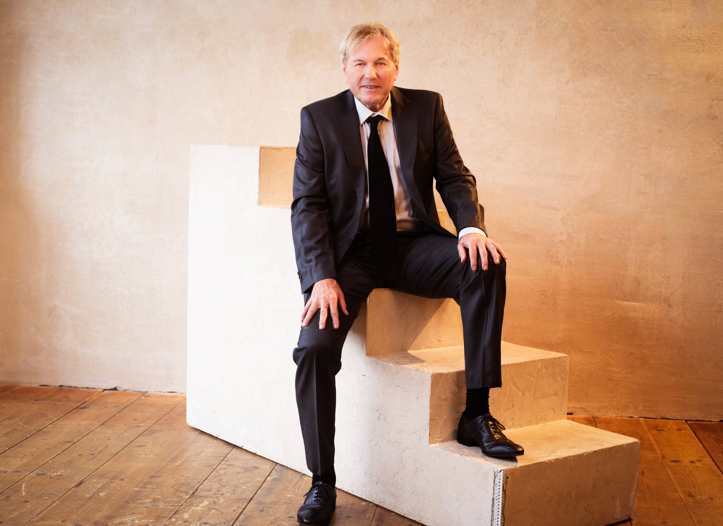 Bernhard Brink in einem schwarzen Anzug, weißem Hemd und schwarzer Krawatte sitzt auf einer Treppe vor einer beigefarbenen Wand.