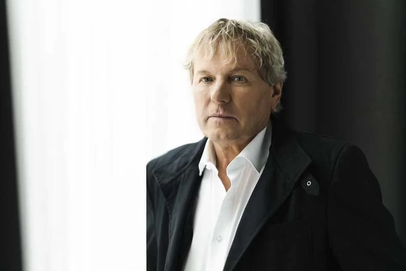 Bernhard Brink mit blondem, leicht verwuschelt Haar, trägt ein weißes Hemd und eine dunkle Jacke, blickt ernst in die Kamera, steht vor einer hellen und dunklen Hintergrundseite.