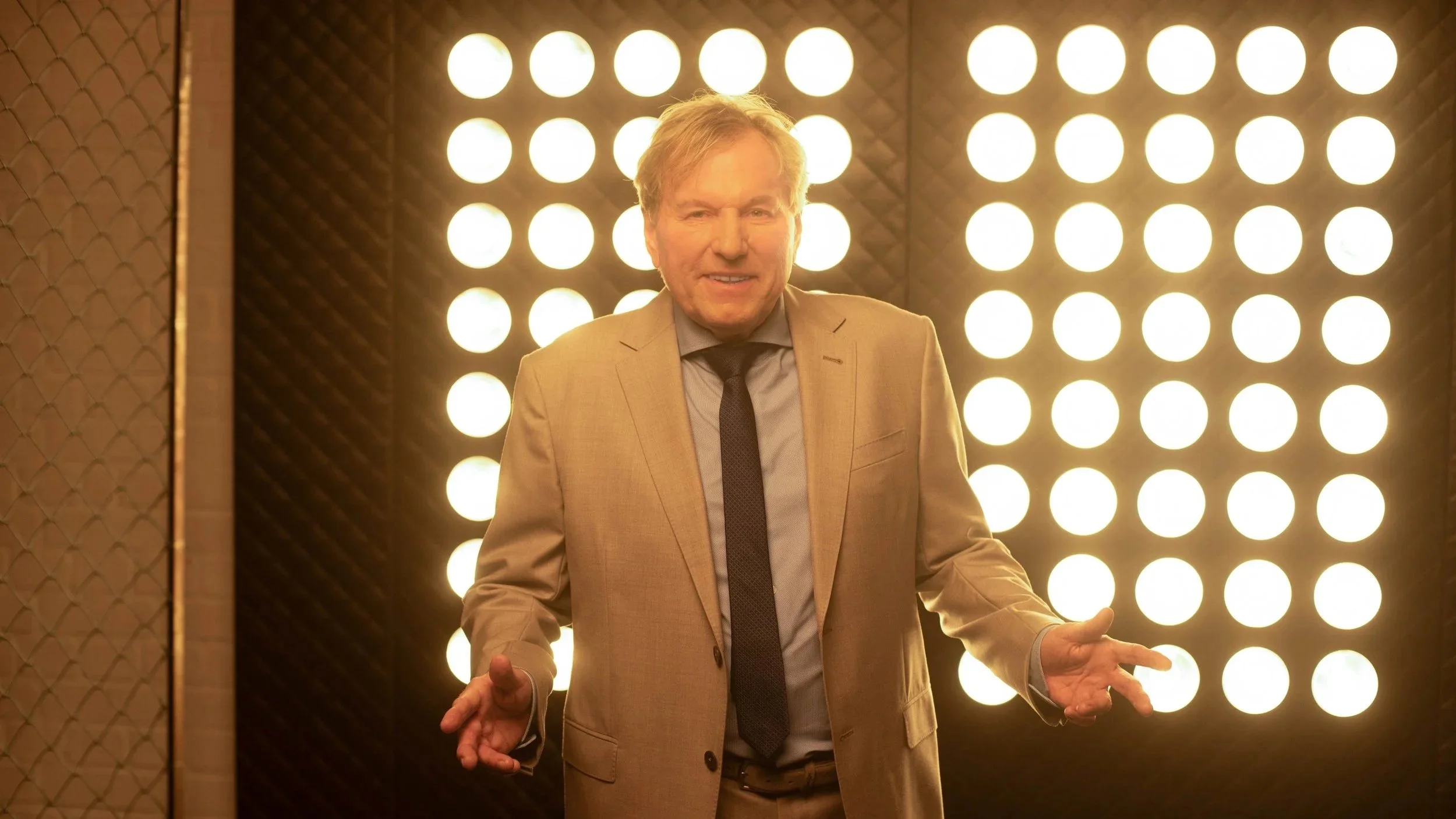 Bernhard Brink in einem beige Anzug und Krawatte steht vor einer Wand mit runden Lichtern, macht eine erklärende Geste.