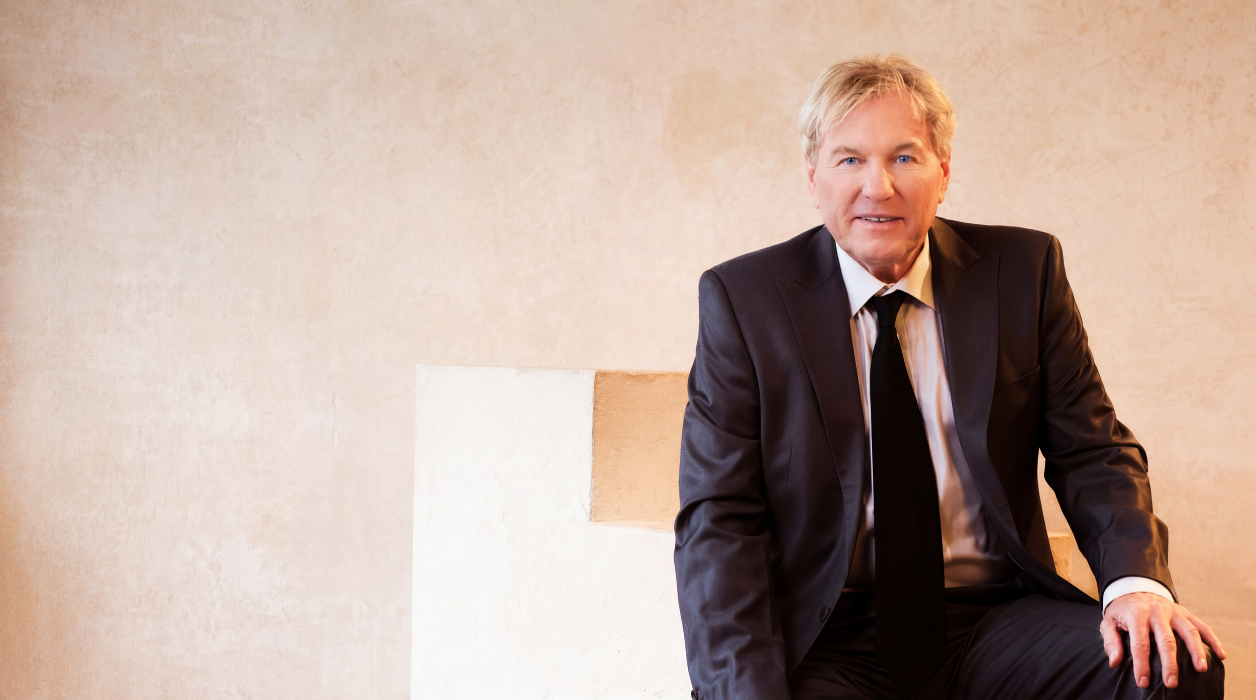 Bernhard Brink in einem schwarzen Anzug sitzt vor einer beige-farbenen Wand und blickt in die Kamera.