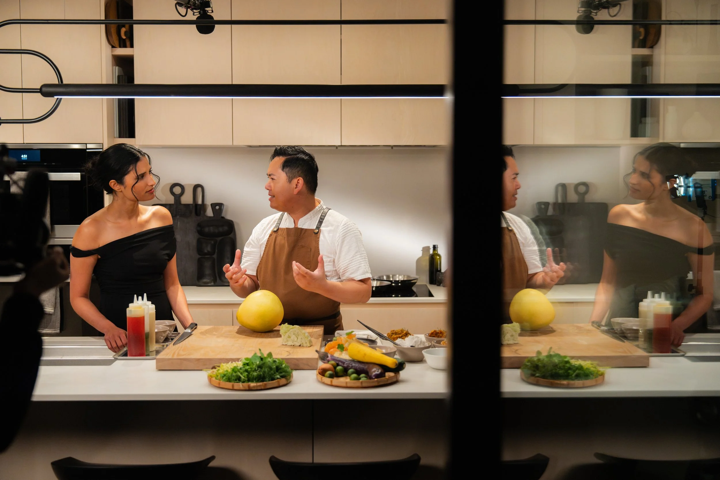 Chef explaining food preparation to a woman in a modern kitchen, with fresh vegetables and cooking ingredients on the counter