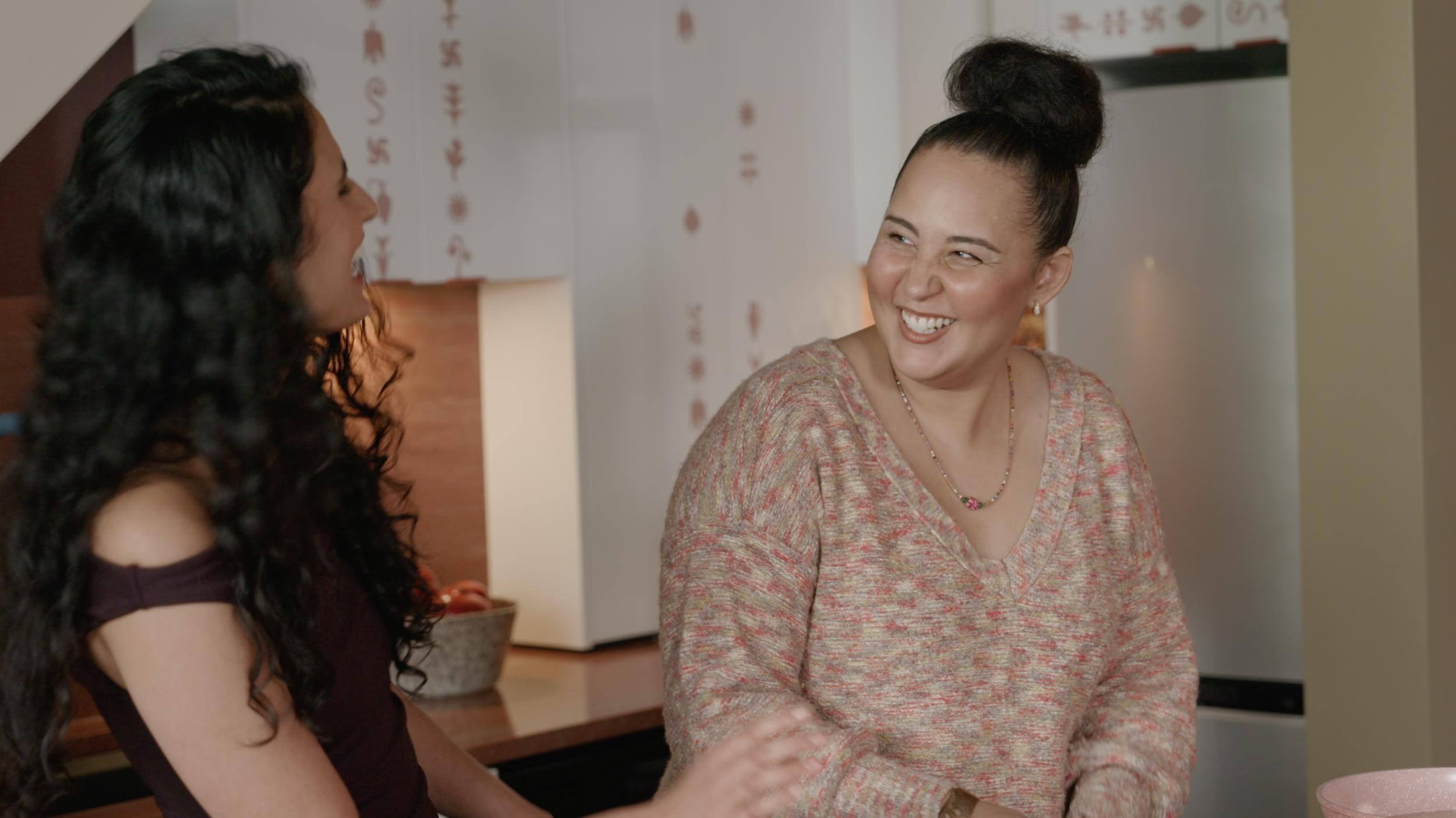 Two women laughing and chatting in a warmly lit kitchen.