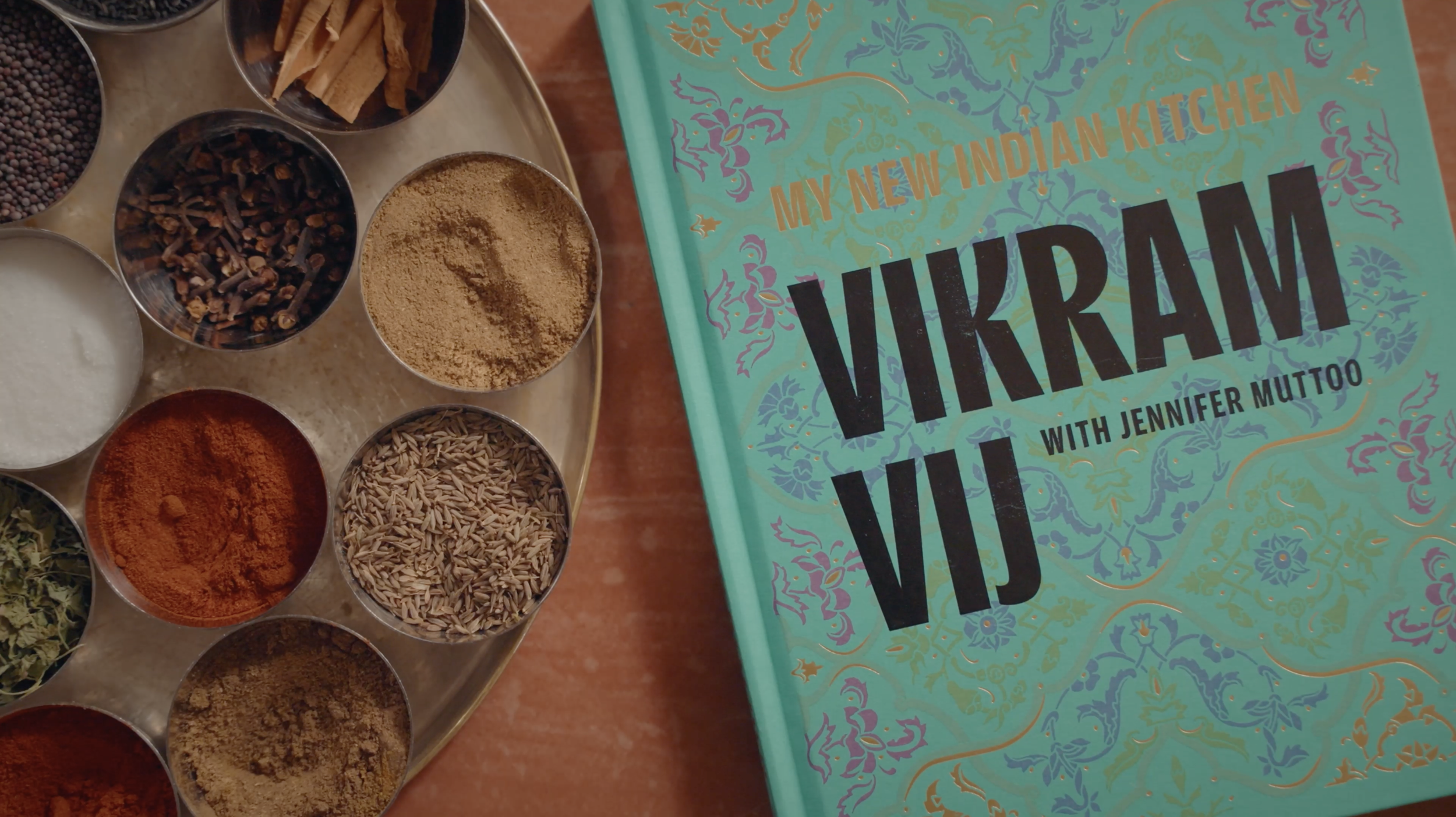 My New Indian Kitchen cookbook beside bowls of colorful Indian spices and whole spices