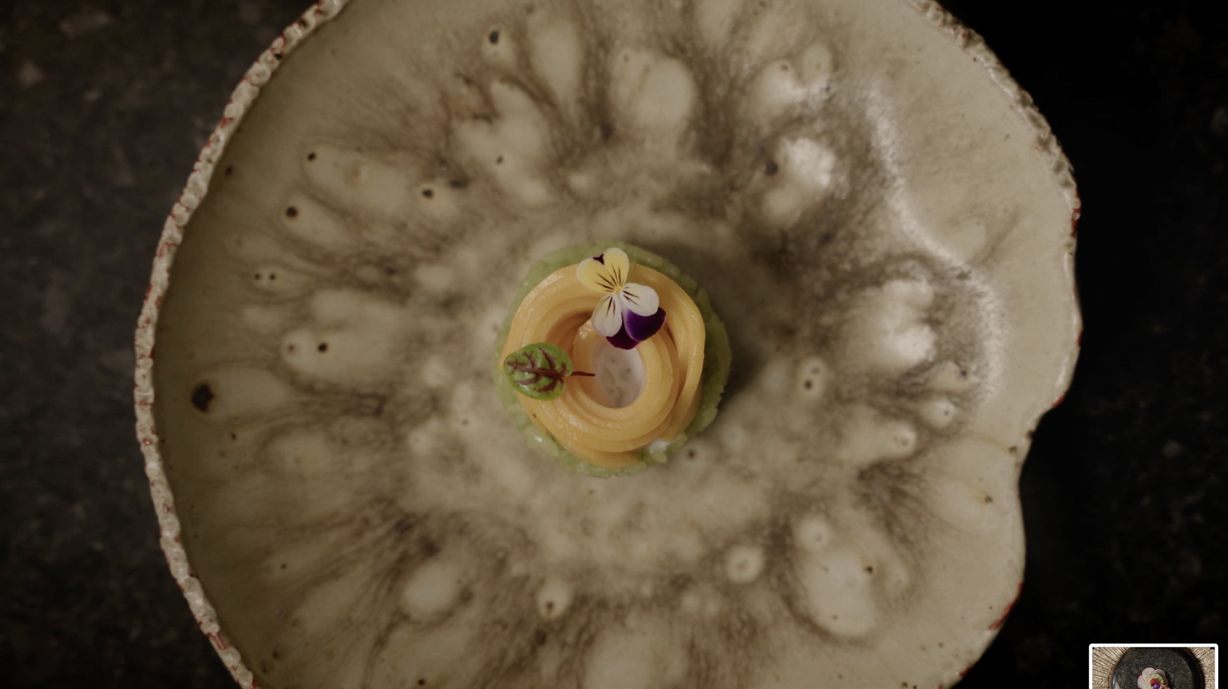 Top-down view of a plated dessert with orange spiral garnish, green accents, and an edible purple-and-white flower on a rustic ceramic plate.