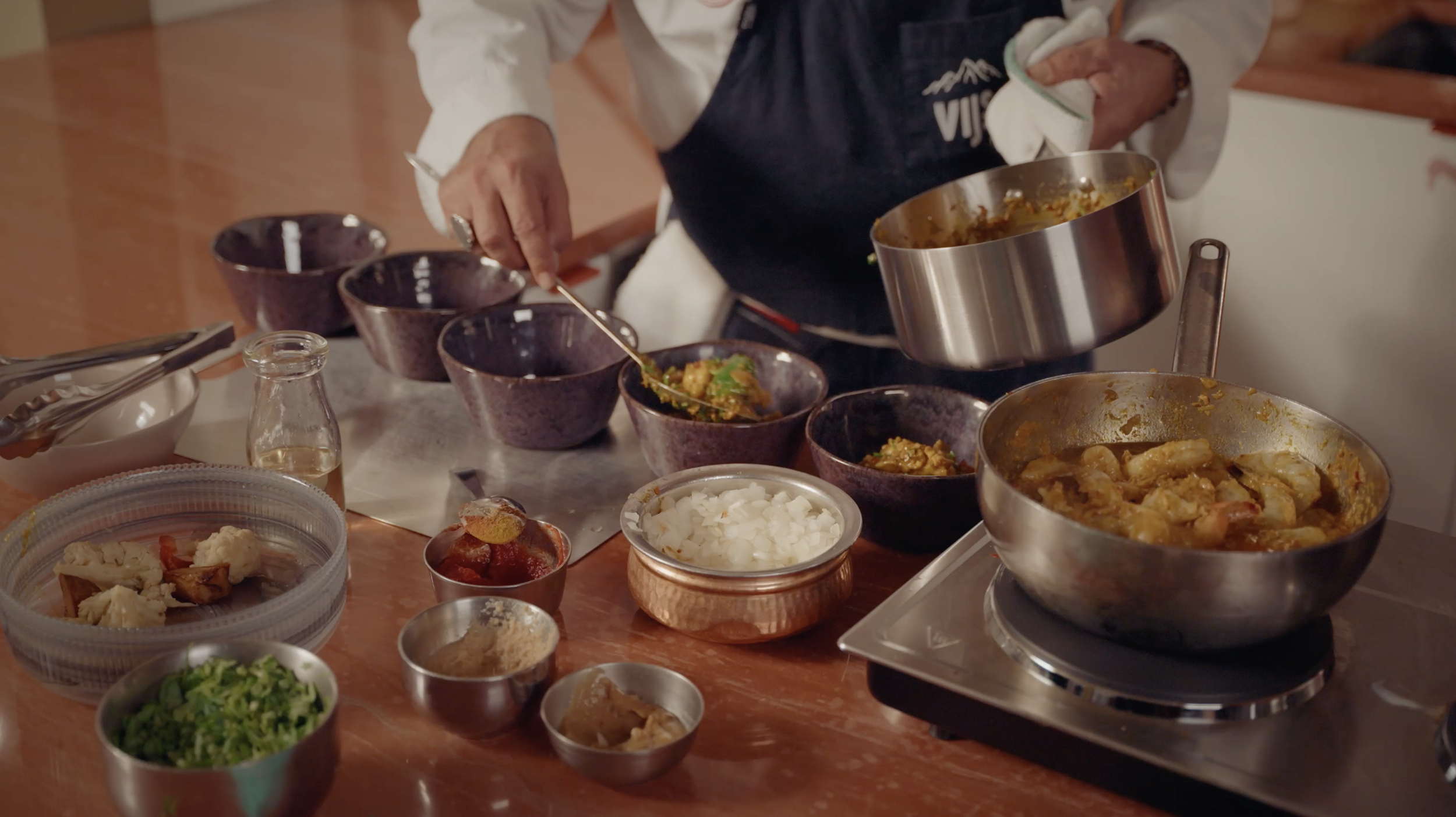 Chef serving Indian food into bowls at a restaurant table with rice, curry, vegetables, and cooking pots.