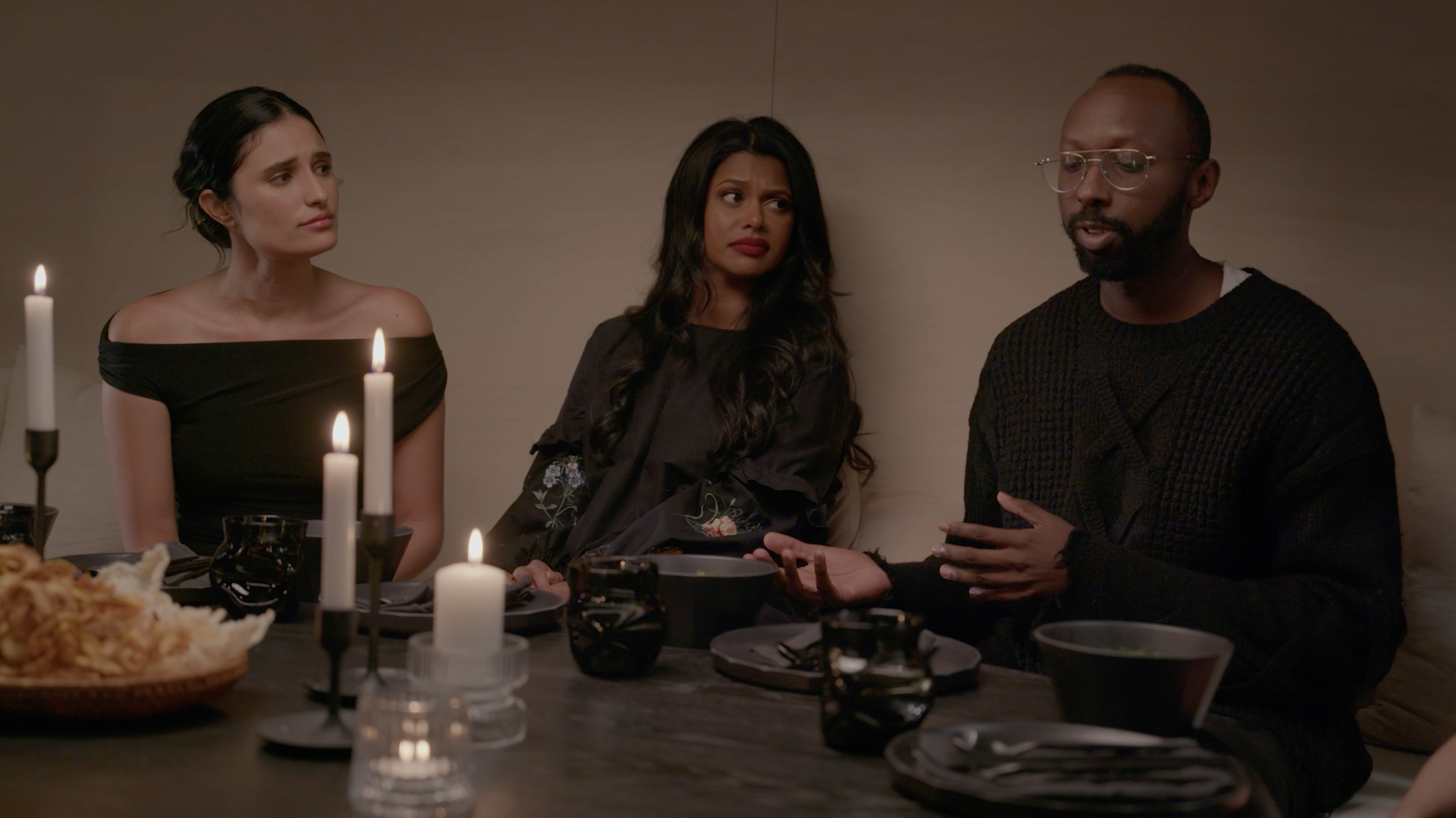 Three people in black clothing sit at a candlelit dinner table, with plates, bowls, wine glasses, and bread.