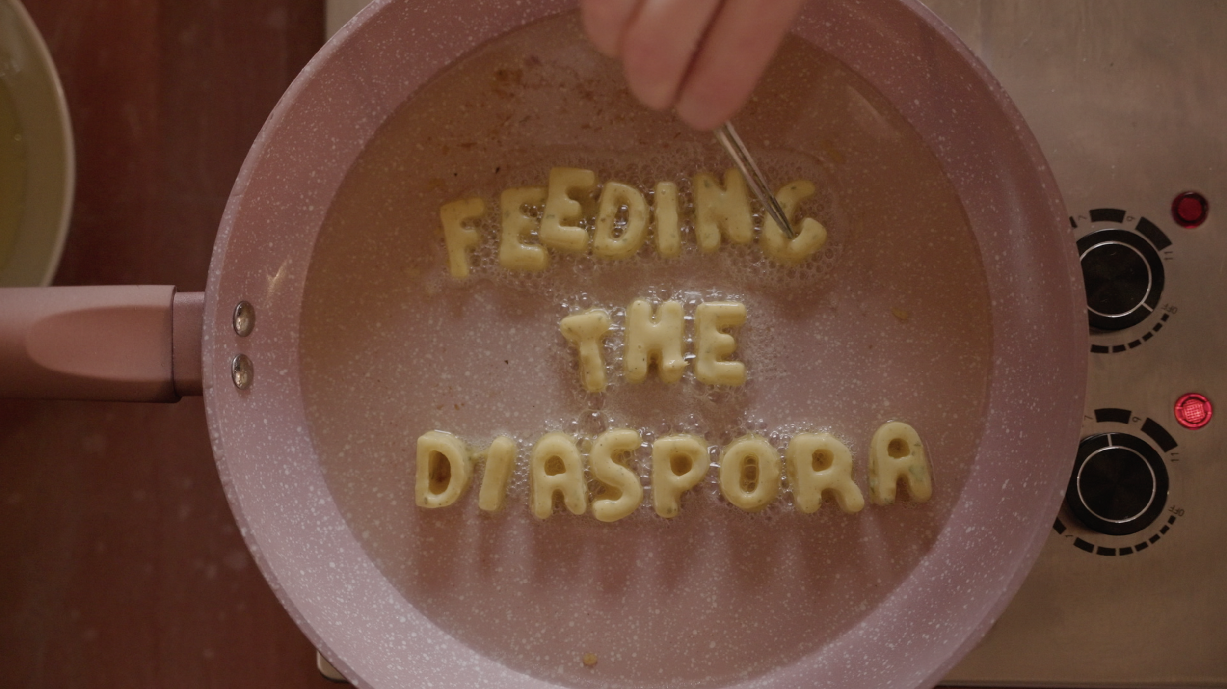 Alphabet pasta spelling “FEELING THE PASPORA” boiling in a pink saucepan on an electric stovetop.