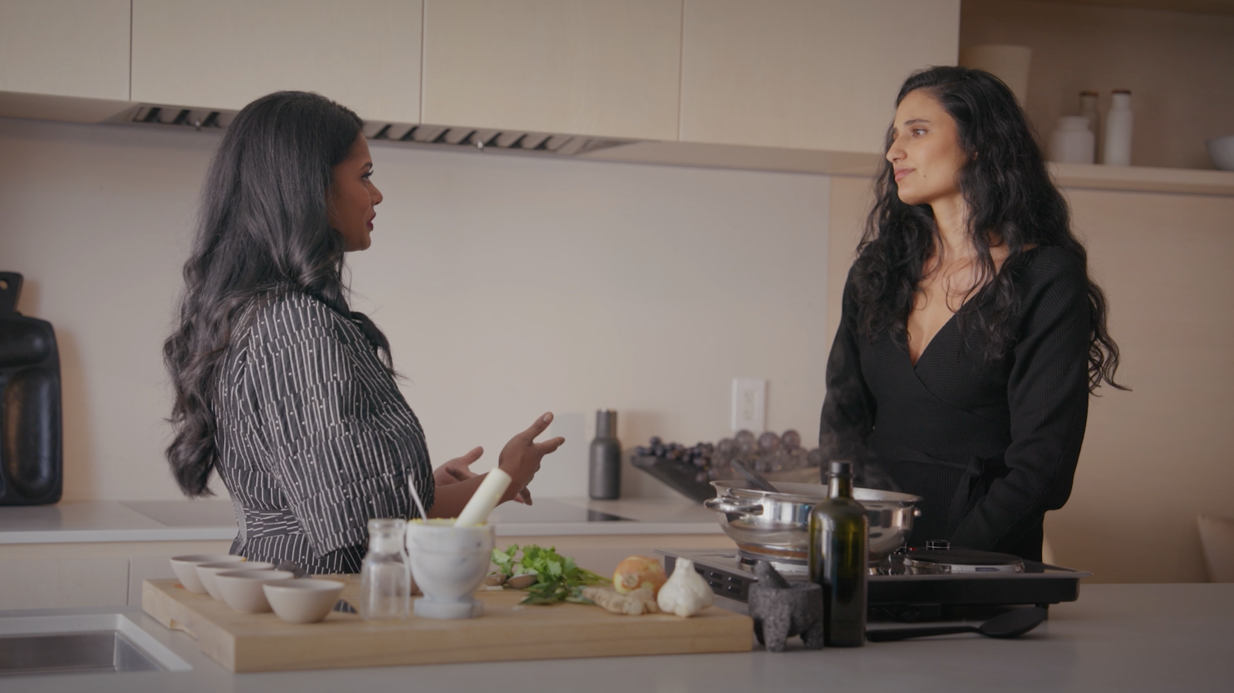 Two women prepare food and talk in a modern kitchen with vegetables, bowls, cookware, and a portable burner.