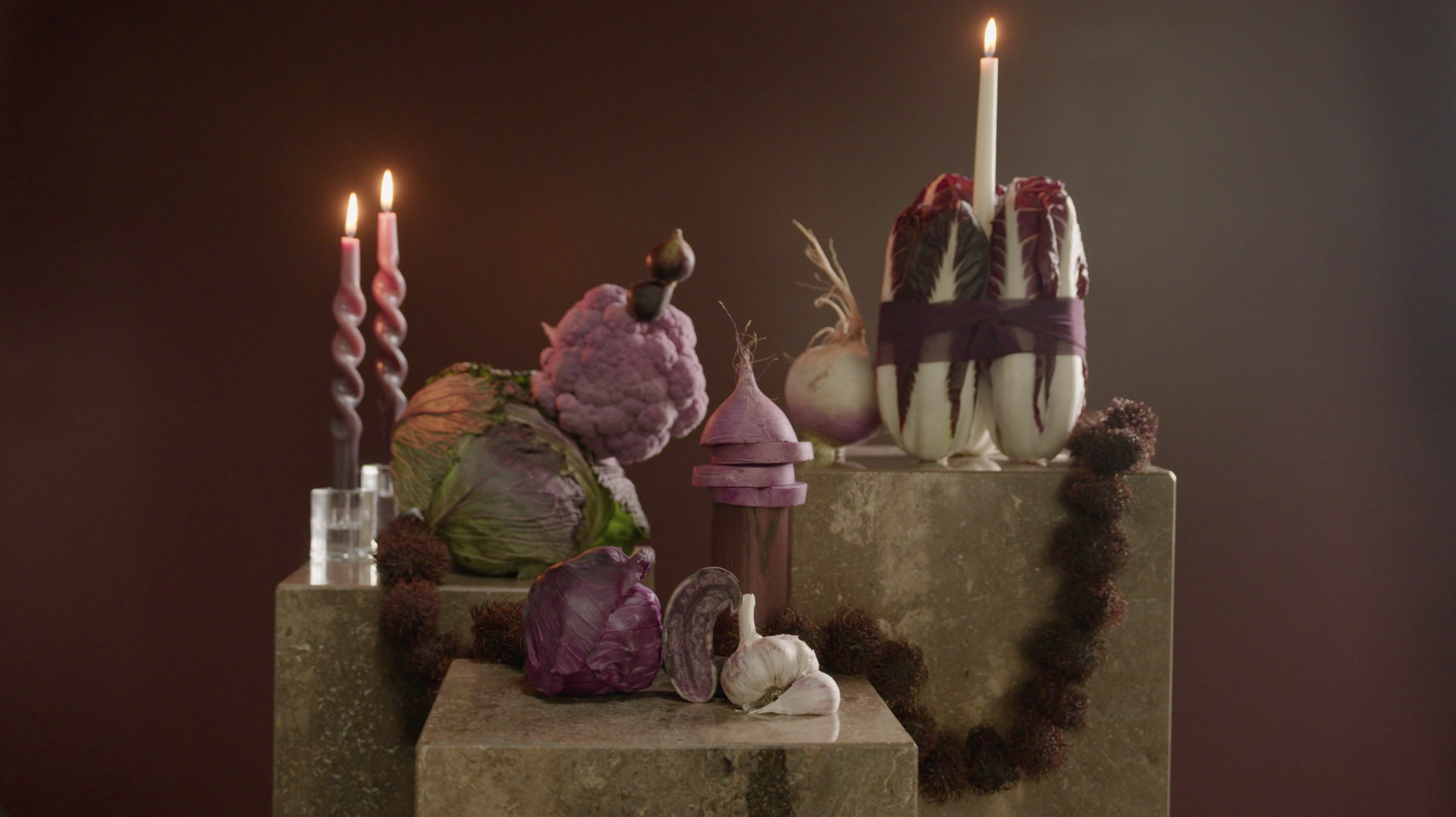 Artistic still life of colorful vegetables, garlic, onions, and candles arranged on stone blocks against a dark background