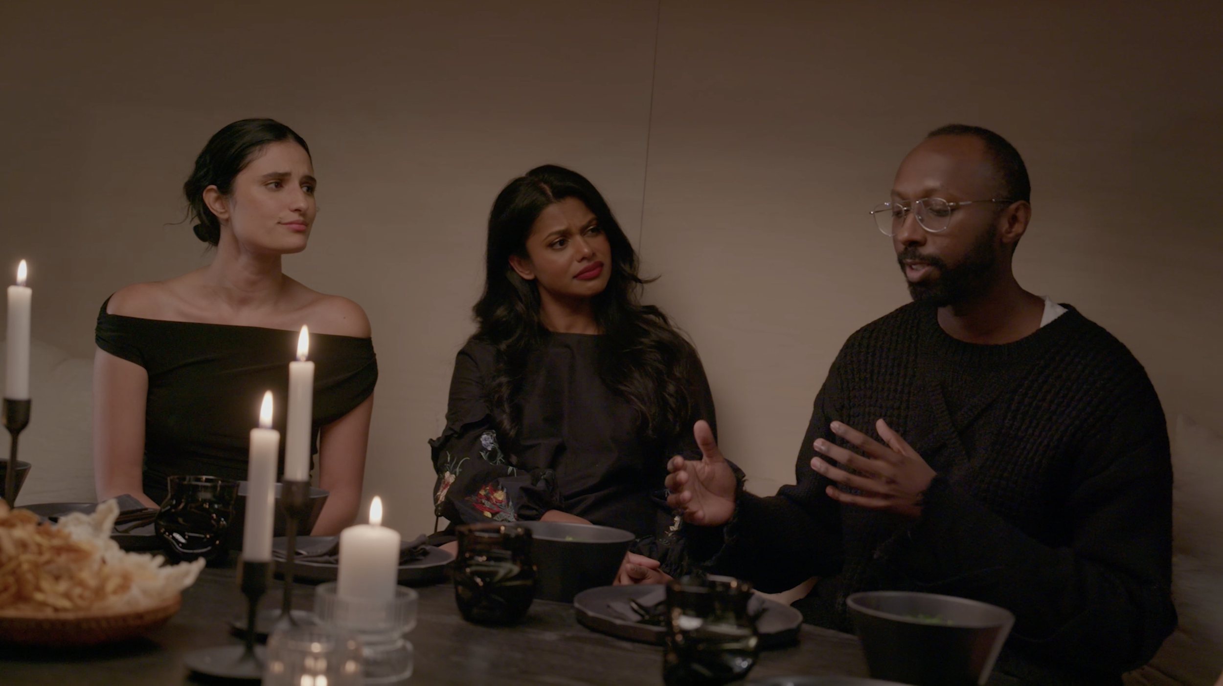 Three people dine at a candlelit table, with a man speaking beside two women.