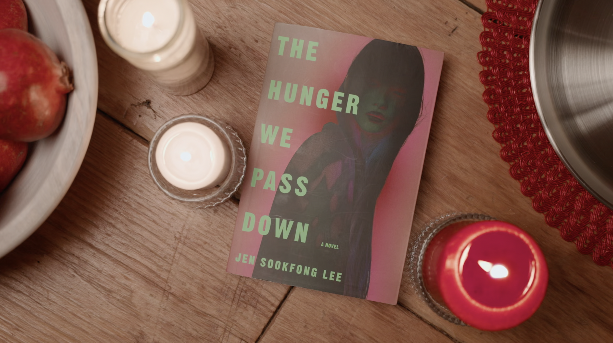 The Hunger We Pass Down by Jen Sookfong Lee on a wooden table with lit candles, pomegranates, and a red placemat.
