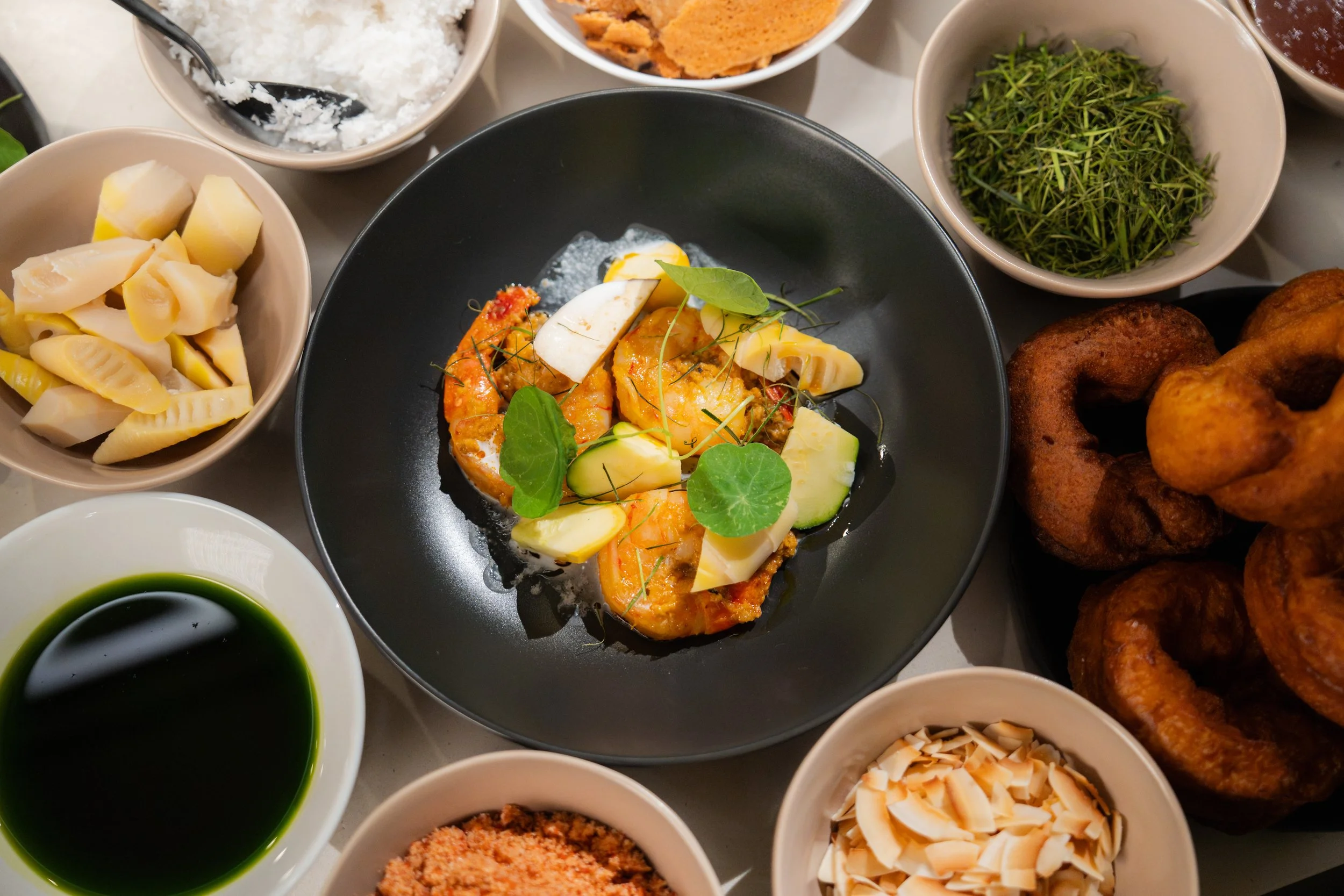 Southeast Asian meal with a plated orange curry, vegetables, herbs, rice, bamboo shoots, fried snacks, sauces, and garnishes