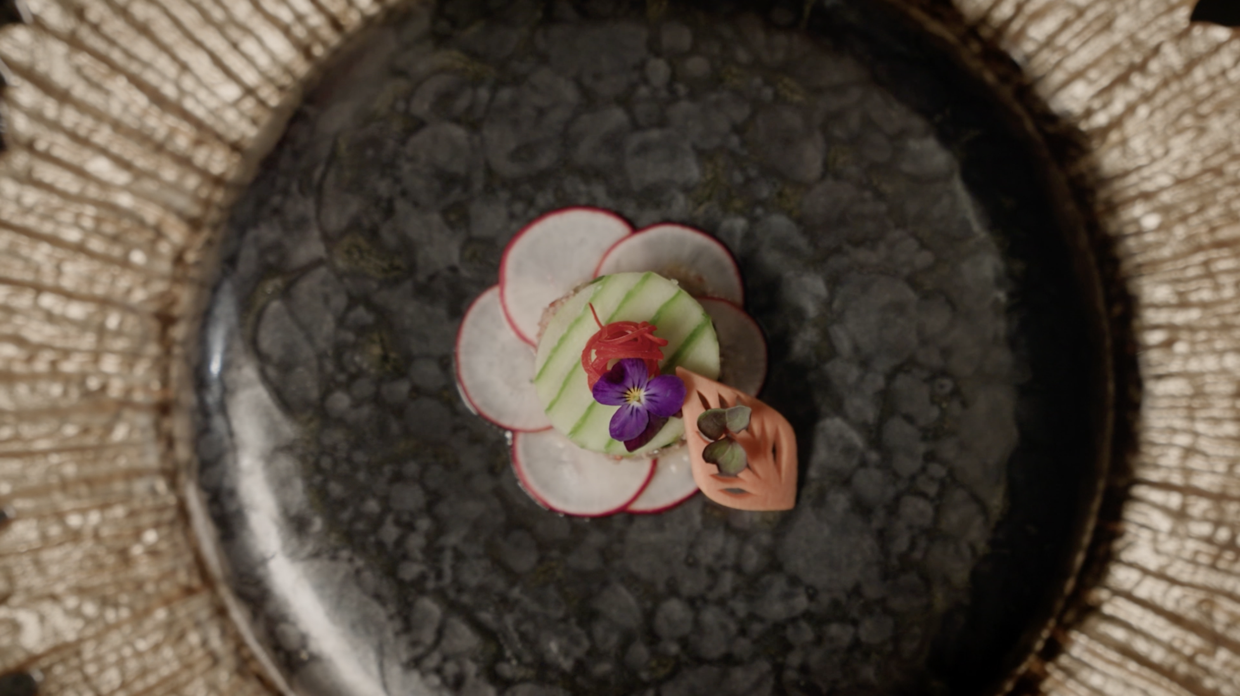 Overhead view of an artistic dish garnished with radish slices, cucumber, pickled ginger, edible purple flower, and greens on a black plate