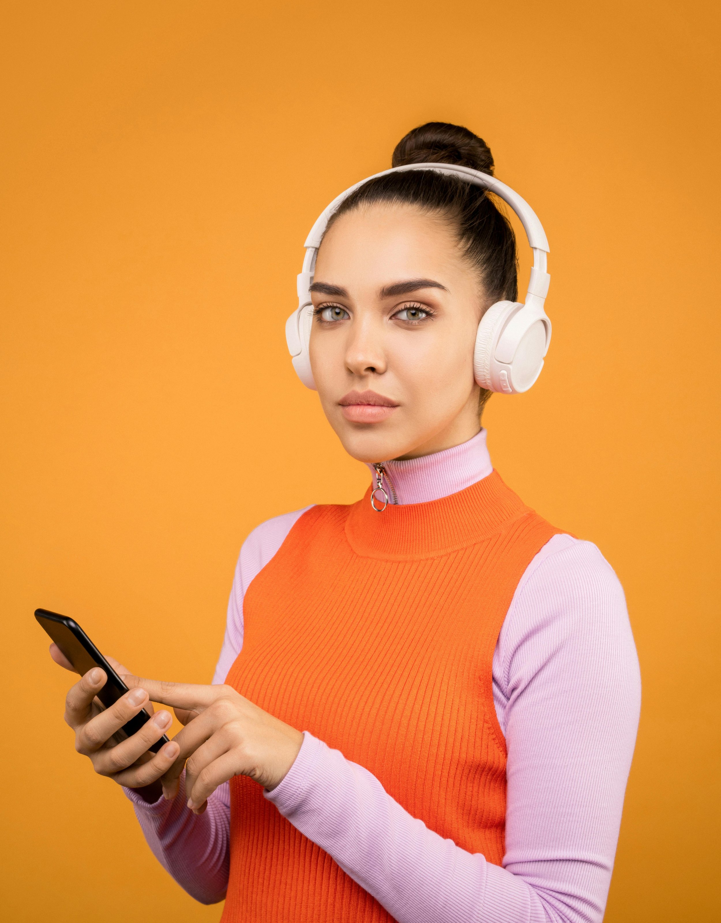 Young woman with dark hair in a bun, wearing white headphones and an orange sleeveless top with pink long sleeves, holding a smartphone against an orange background.