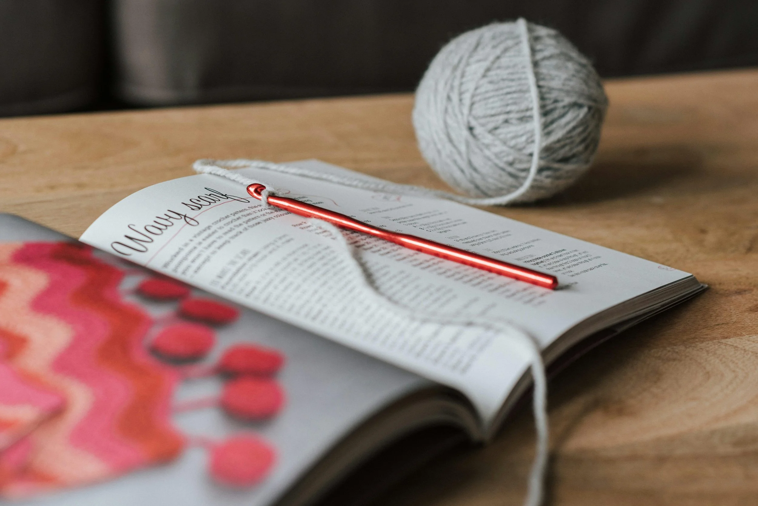 An open magazine or catalog on a wooden table, a red crochet hook lying on top, a ball of yarn in the background, and a colorful crochet project partially visible in the foreground.