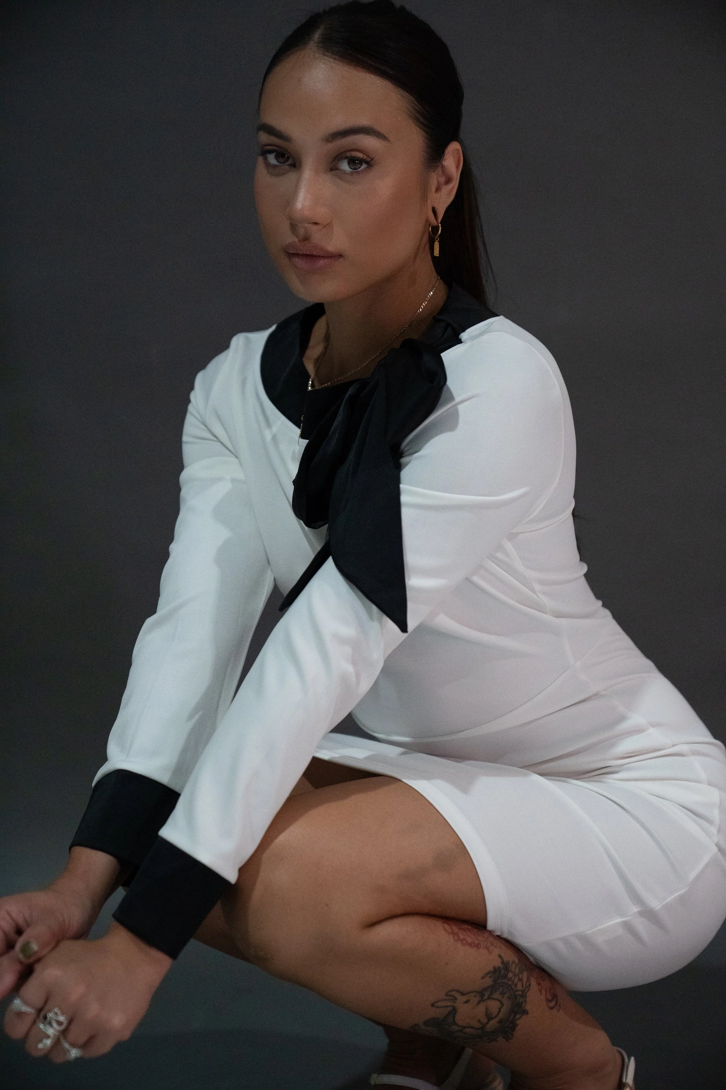 A woman with dark hair, wearing a white dress with black accents and a black scarf, squatting against a dark background.