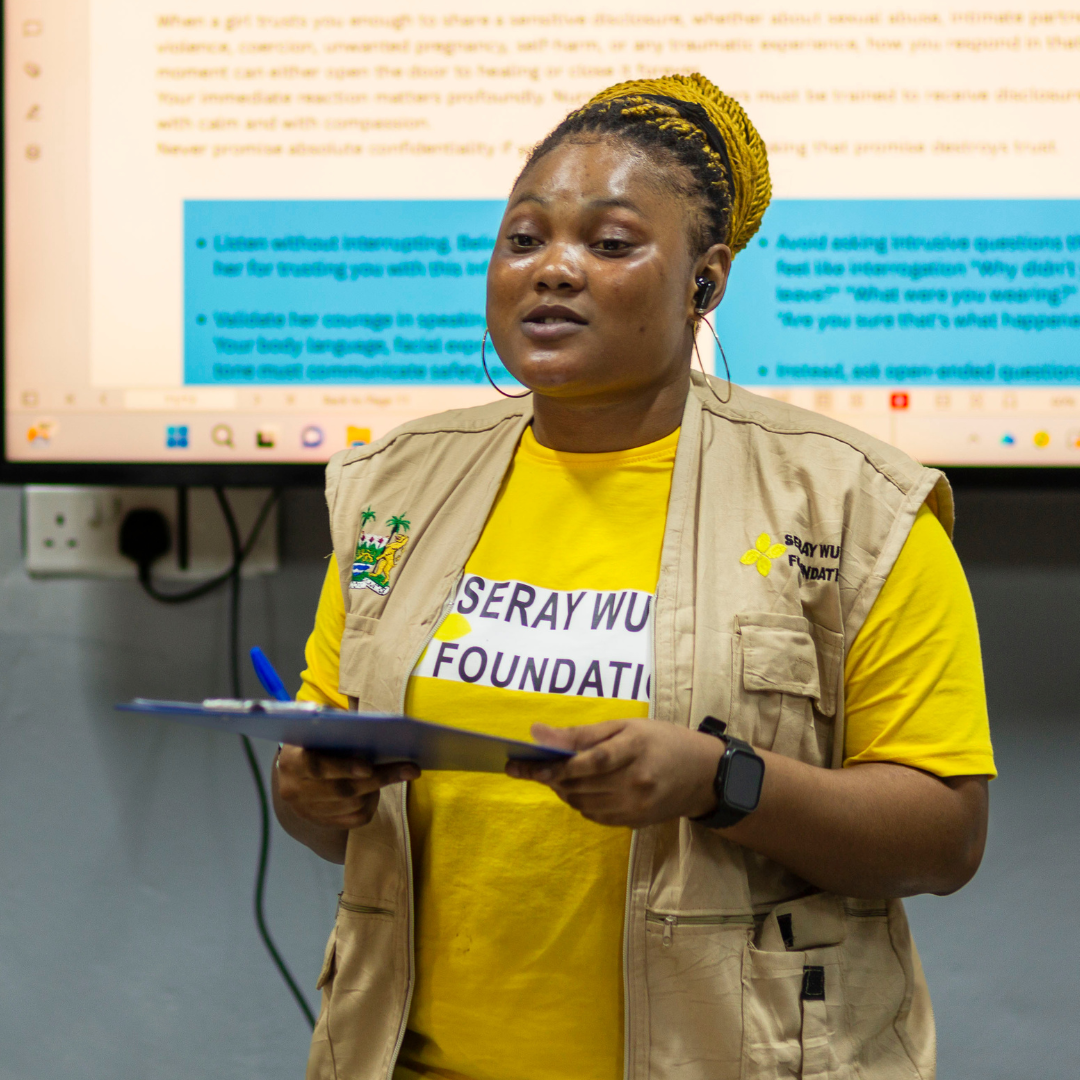 A woman in a yellow shirt and beige vest speaking in front of a large screen during a presentation, holding a clipboard, with a focused expression.