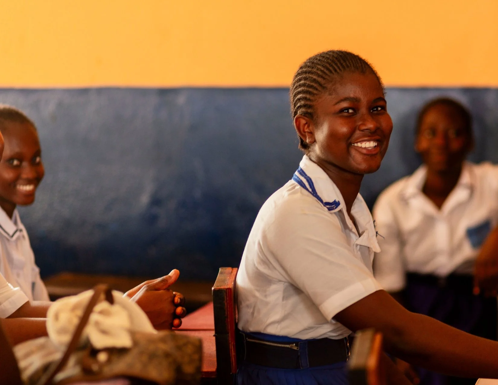 Smiling schoolgirl with braided hair wearing a white school uniform with blue stripes, sitting at a table in a classroom.