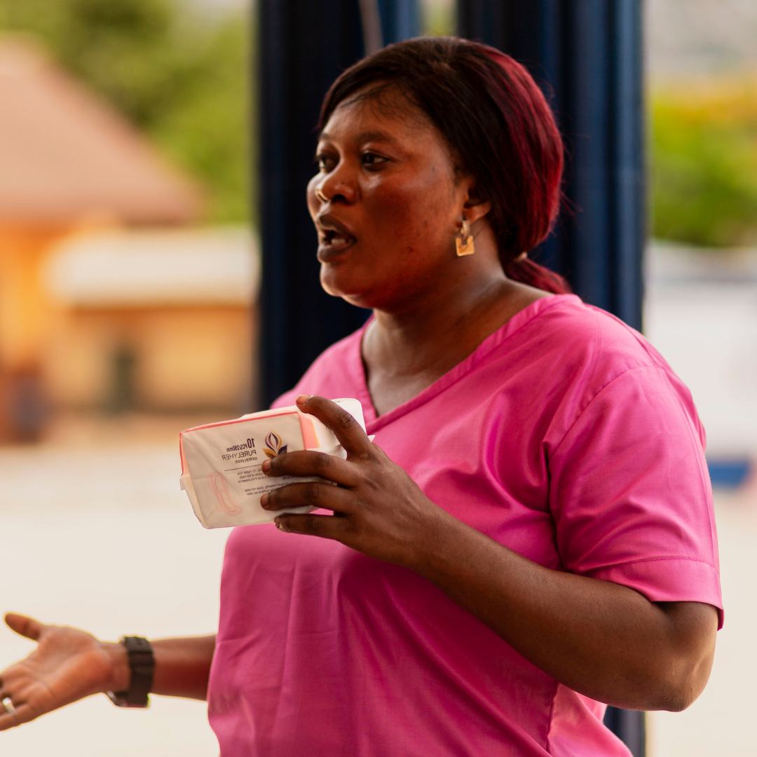 A woman with shoulder-length dark hair, wearing gold earrings and a pink shirt, holding a white and pink box of PureLife tissue paper, gesturing with her left hand while speaking.
