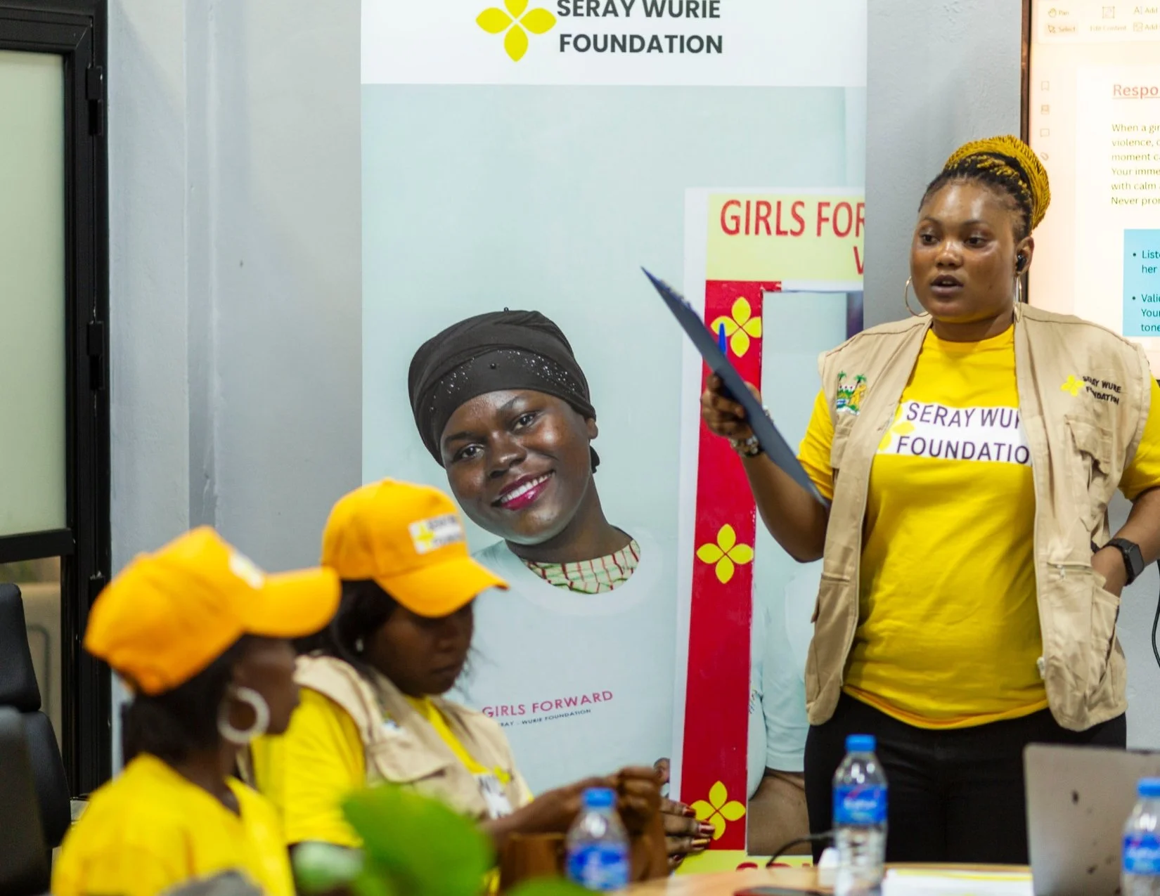 Women wearing yellow shirts and hats with 'Seray Wurie Foundation' logos attending a meeting or event, with a woman standing and speaking, and a poster with the foundation's name in the background.