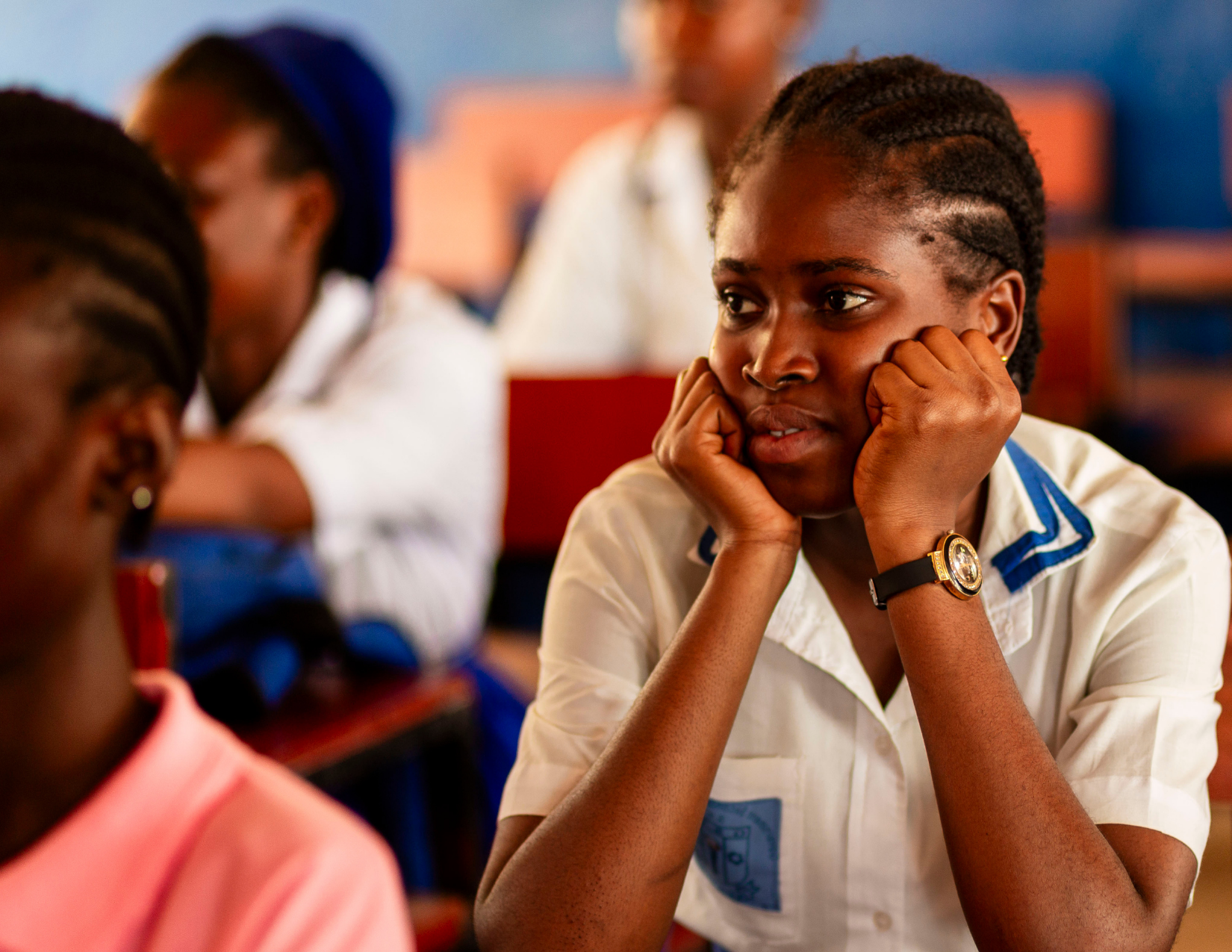 A young African girl in a school uniform is sitting at a desk in a classroom, with her head resting on her hands, looking thoughtful or bored. Other students are visible in the background.