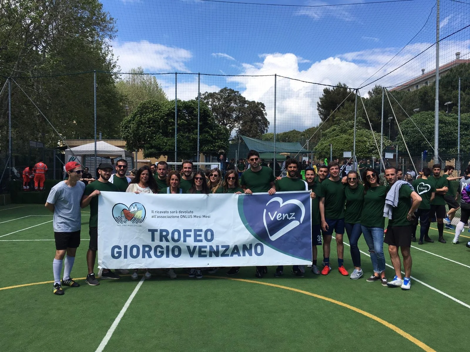Gruppo di persone che partecipano a un evento di beneficenza su un campo di calcio, con un grande striscione che dice 'Trofeo Giorgio Venzano' e 'Il ricavato sarà devoluto all'associazione ONLUS Mesi Mesi'.