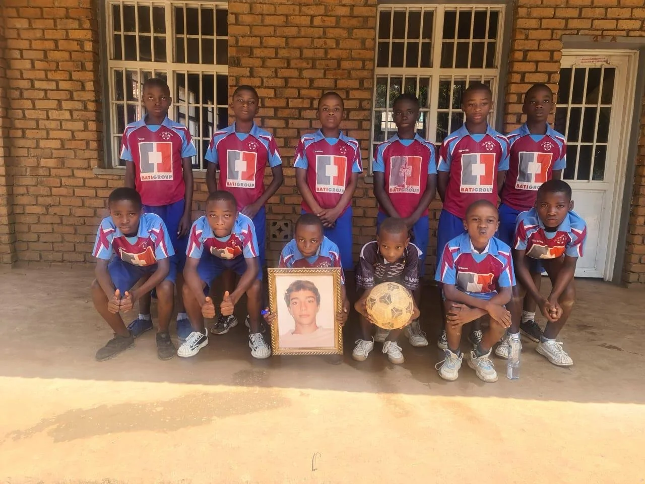 Gruppo di bambini in divisa da calcio con alcuni che tengono una foto di un ragazzo e un pallone, in piedi davanti a un muro di mattoni.