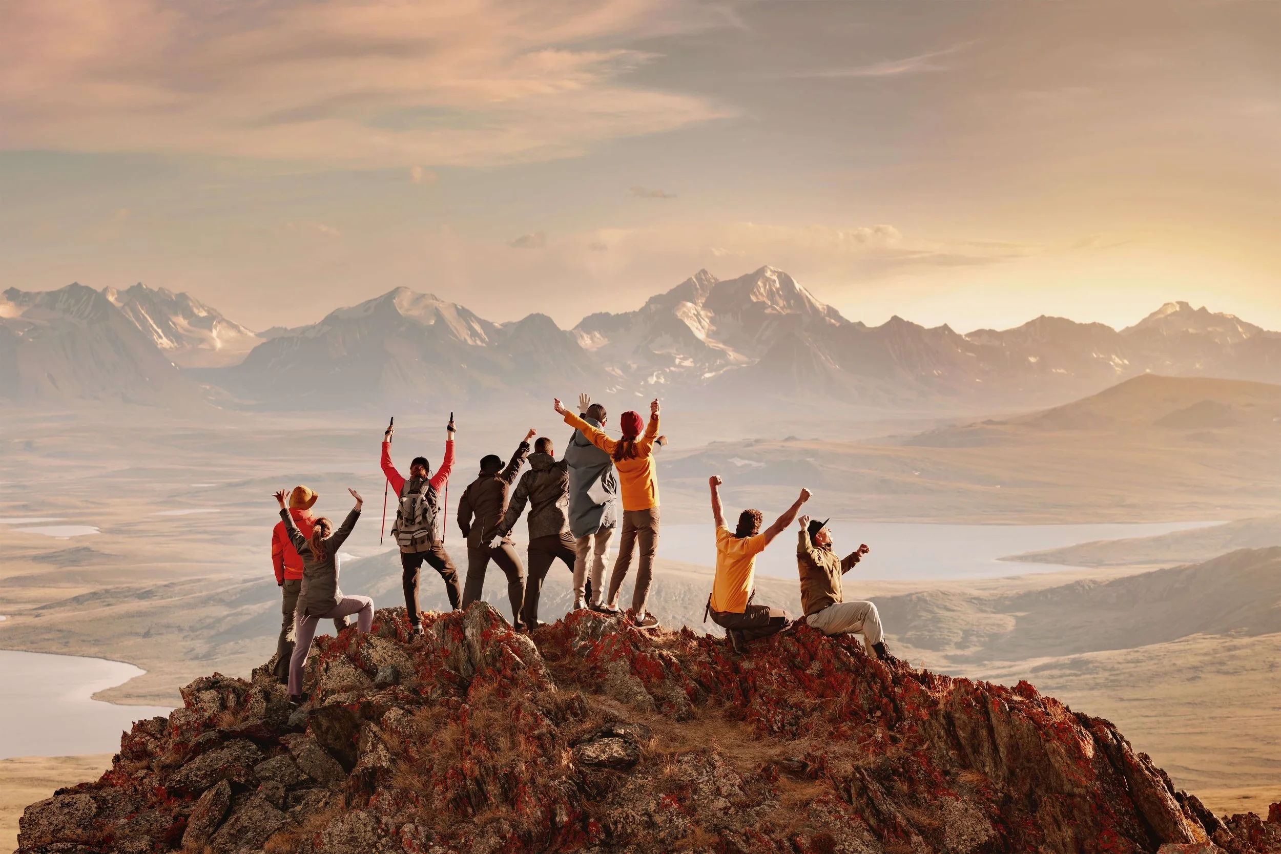 Group of hikers celebrating on a mountain peak with a scenic valley and snow-capped mountains in the background during sunset.