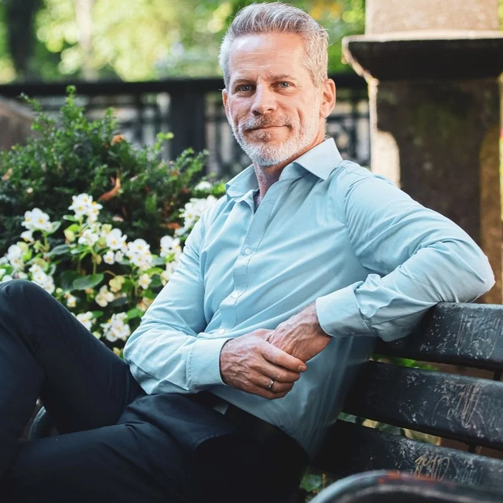 A mature man with gray hair and a beard sitting on a park bench outdoors, wearing a light blue dress shirt and dark pants, with greenery and white flowers in the background.