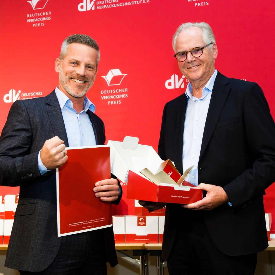 Two men in suits celebrating at an award event with red background and boxes.