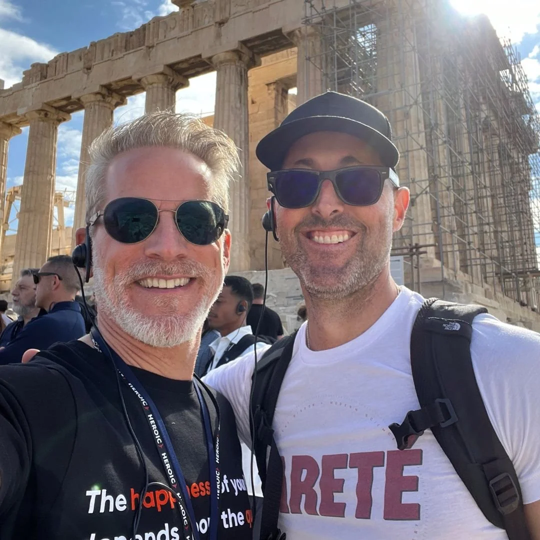 Fabian Erlhöfer und Brian Johnson (heroic), die vor dem Parthenon auf der Akropolis in Athen stehen, bei sonnigem Wetter, tragen Sonnenbrillen und lächeln in die Kamera.