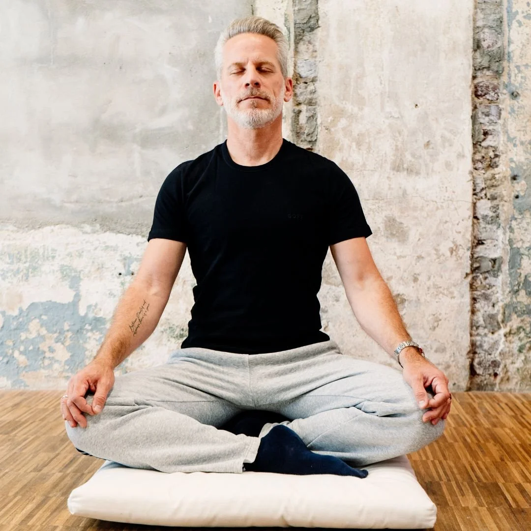 A middle-aged man with gray hair and beard sitting cross-legged on a cushion, practicing meditation with closed eyes against a rustic wall.