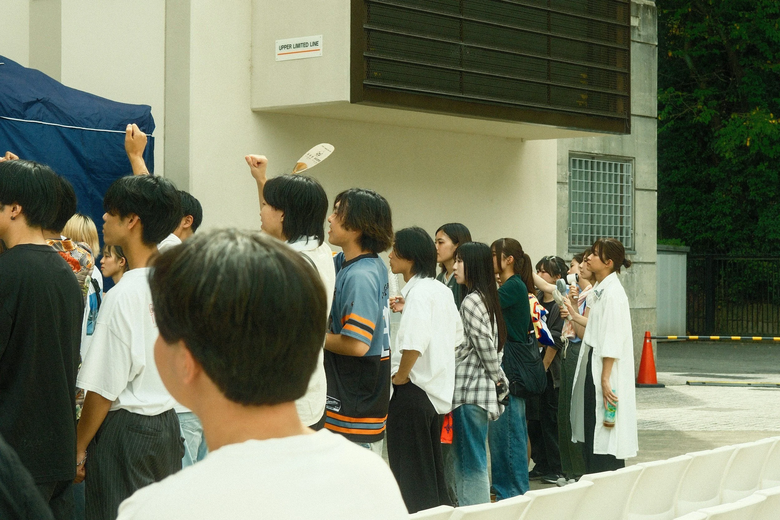 Group of people standing in line outdoors near a building, some raising fists, others holding microphones and water bottles.
